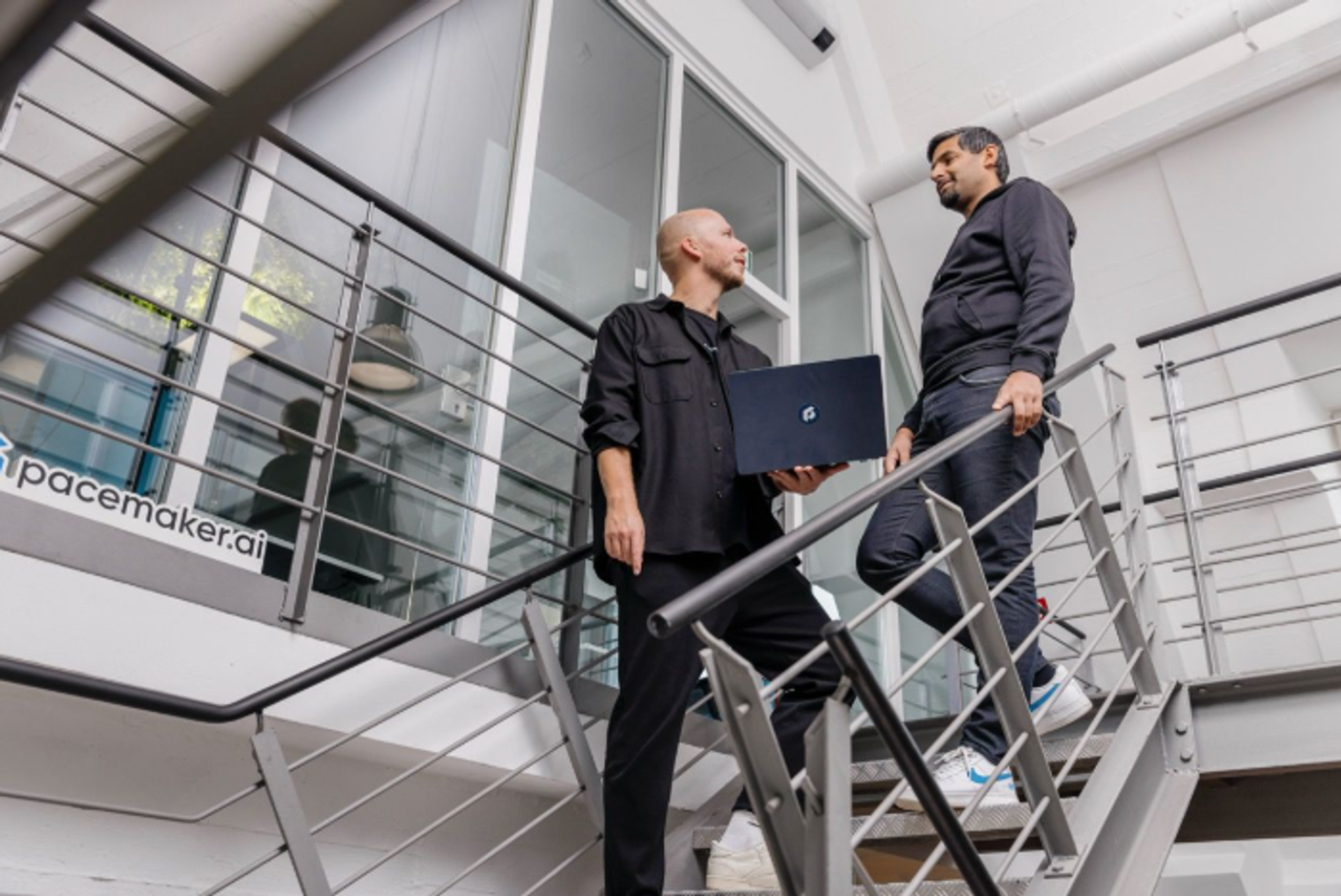 Zwei Mitarbeitende von pacemaker.ai stehen auf einer Treppe im Bürogebäude und sprechen über ein Projekt, einer hält einen Laptop in der Hand.