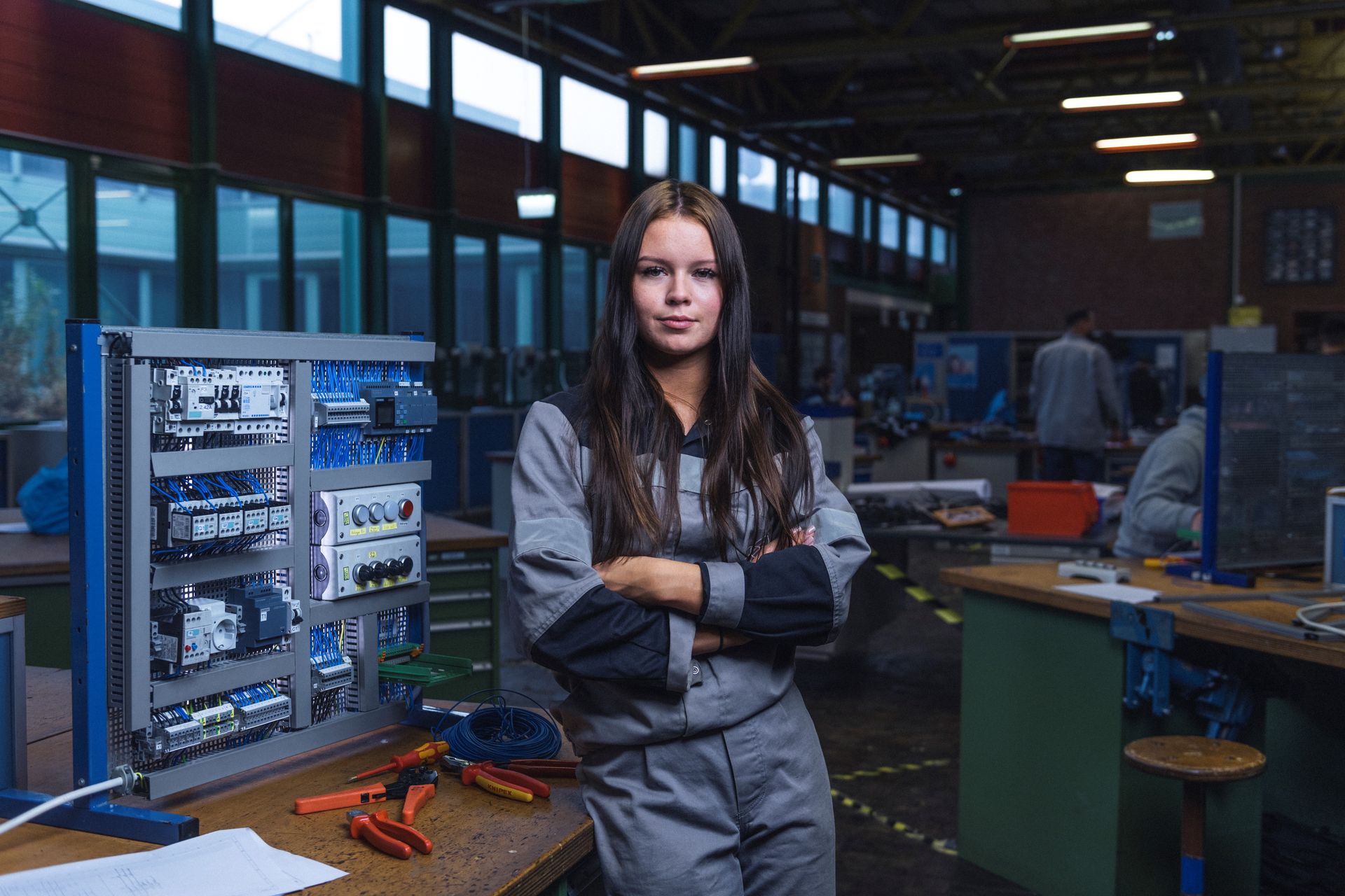 Mädchen lehnt an Werkbank neben offenem Schaltschrank mit Werkzeugen in einer Werkstatt.