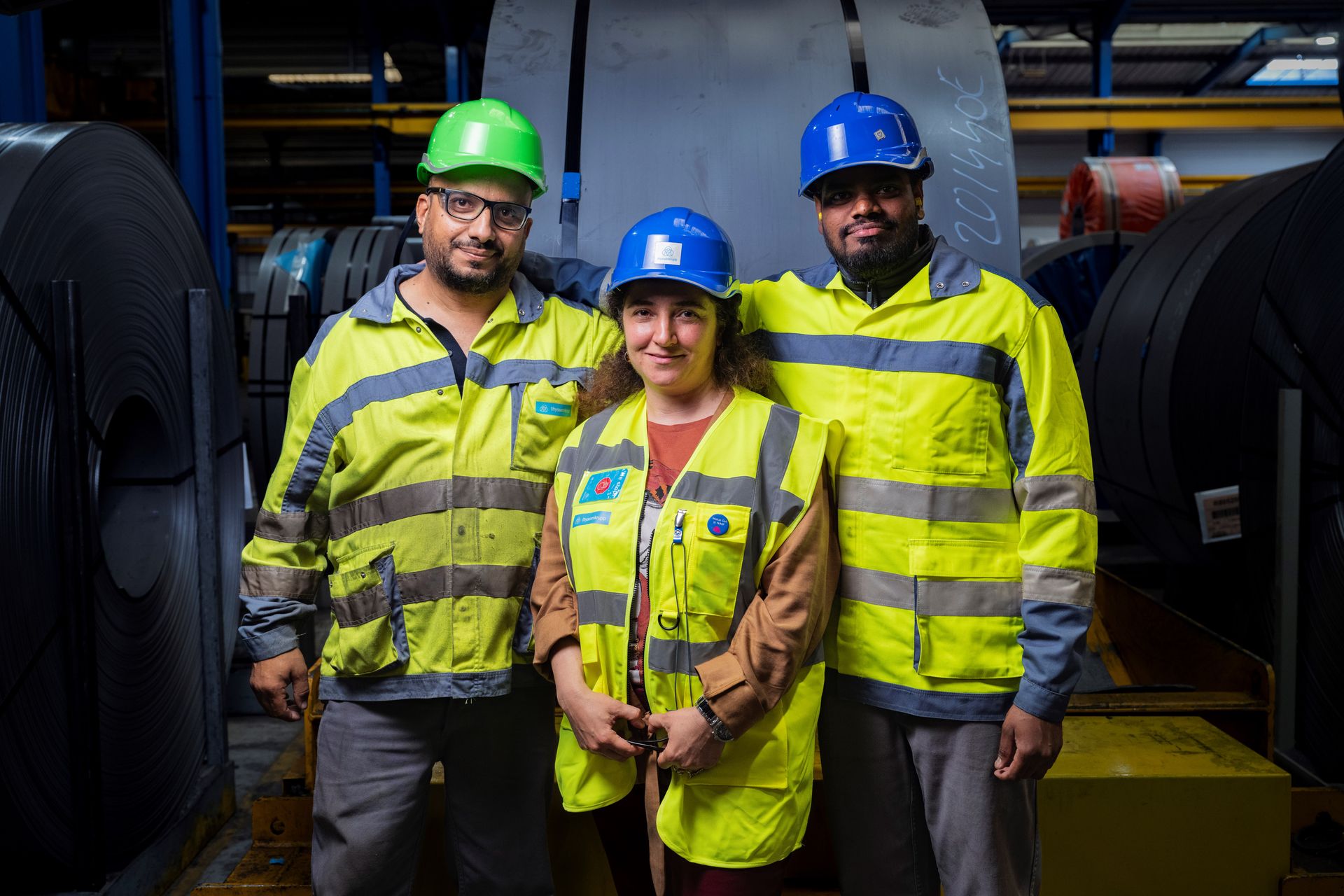 Drei Personen in Sicherheitswesten und Schutzhelmen stehen in einer Industriehalle zwischen großen Stahlrollen.