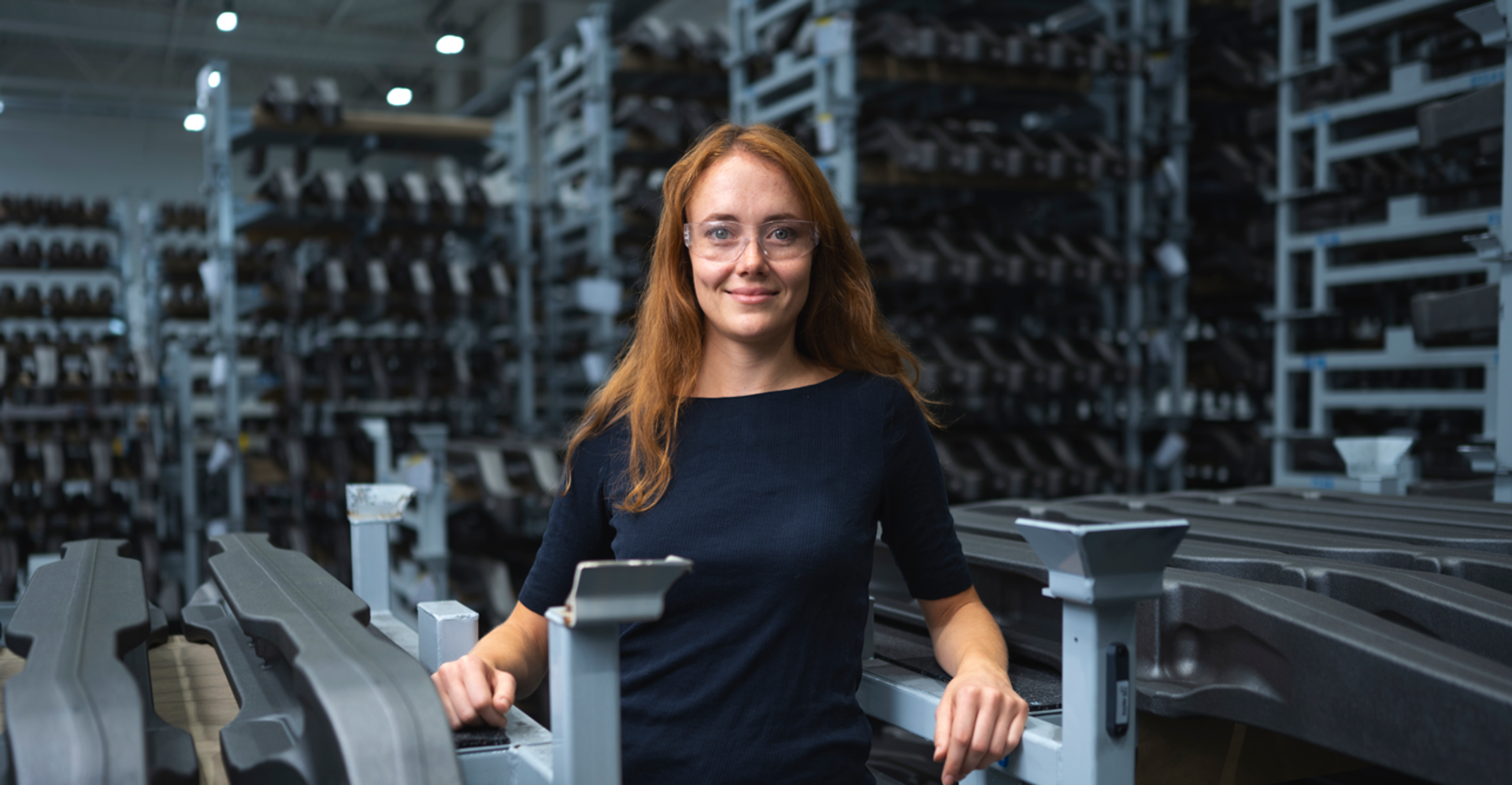Eine Mitarbeiterin steht in einer Produktionshalle zwischen metallischen Bauteilen und Industriegestellen. Sie trägt Schutzbrille und lächelt selbstbewusst in die Kamera.