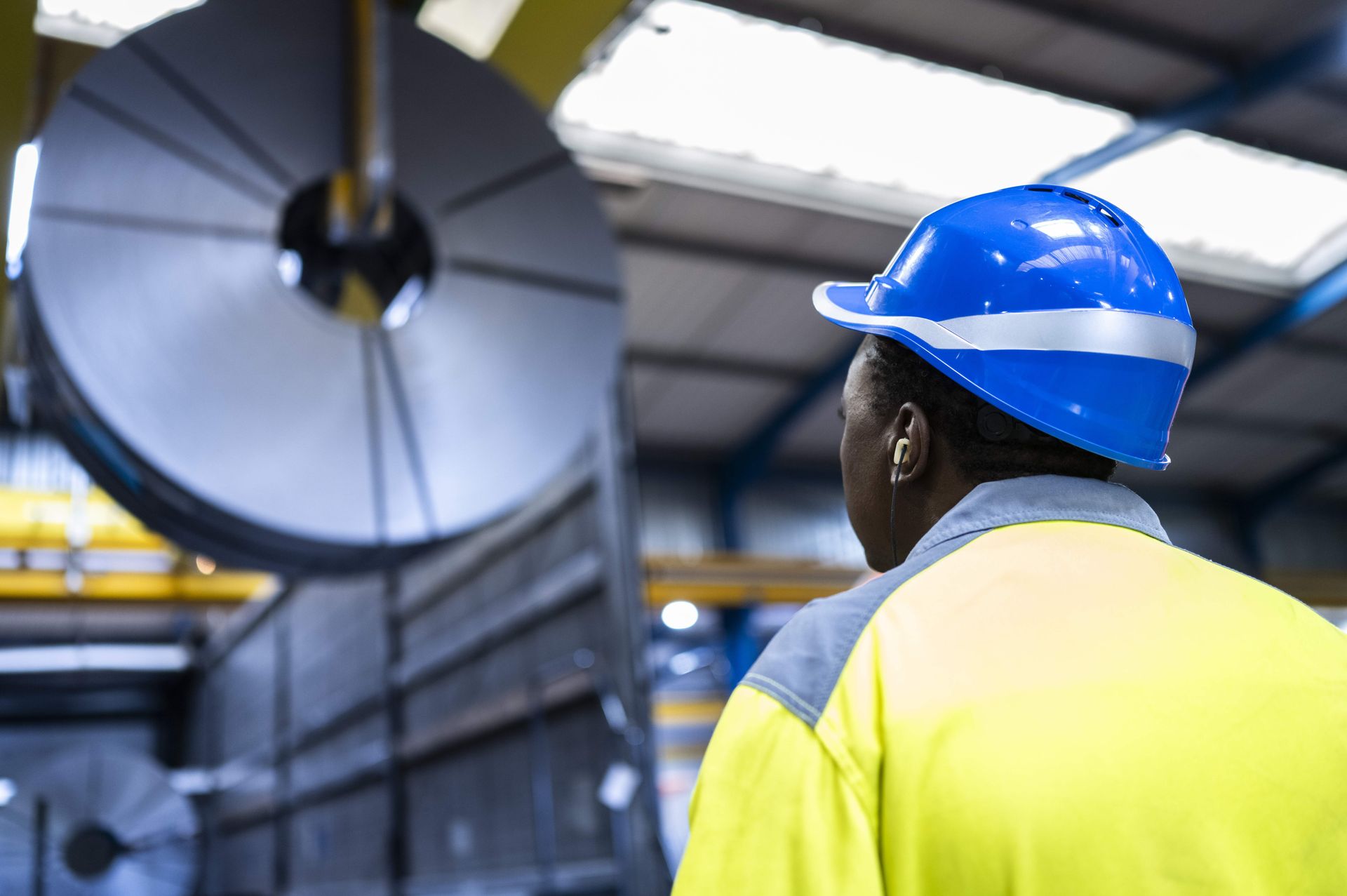 Ein Mitarbeiter mit Schutzhelm und Warnkleidung steht vor einem großen Stahlcoil in einer Industriehalle.