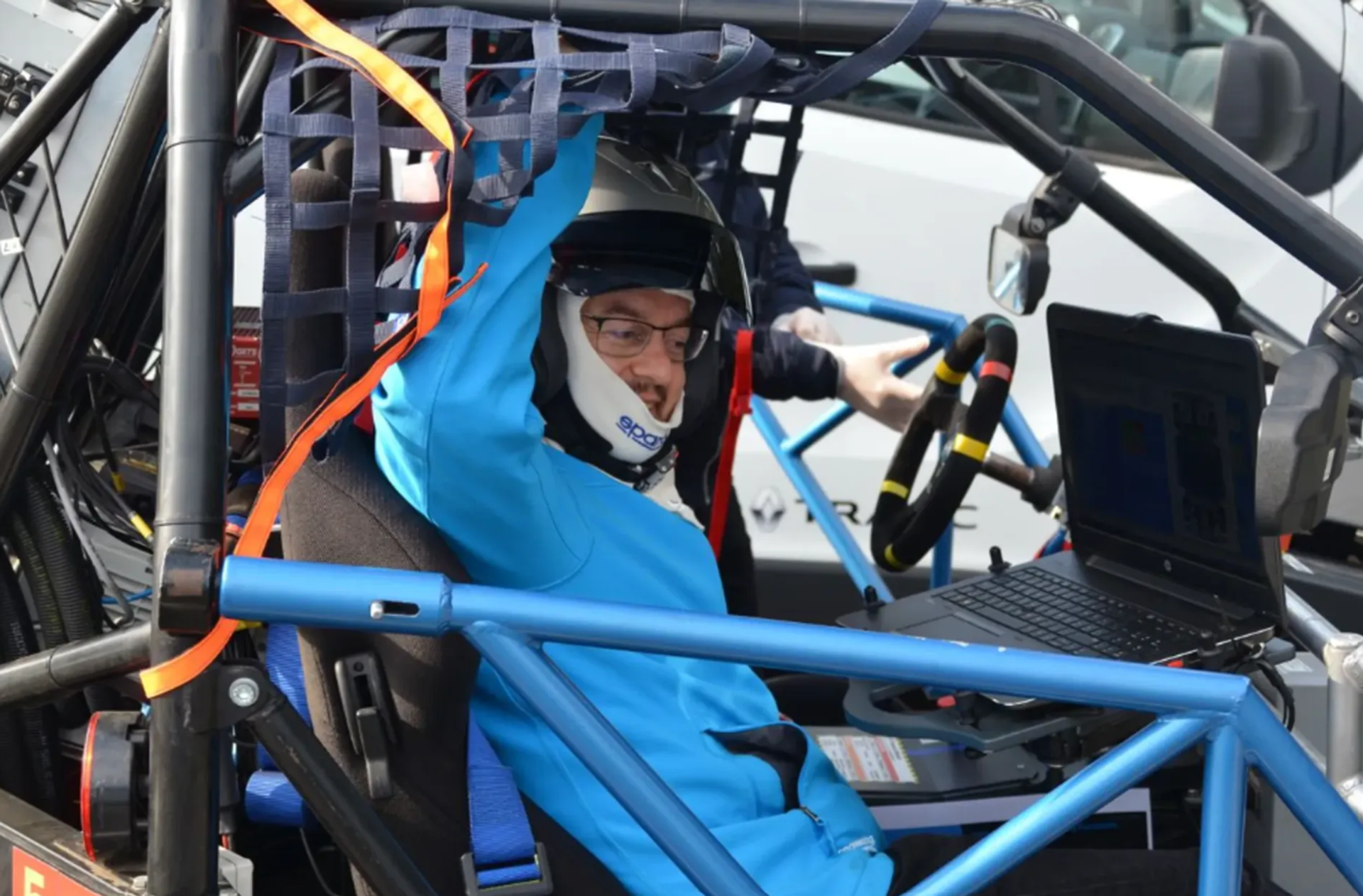 Male-presenting person wearing a helmet sits in a vehicle prototype with a roll cage; a laptop is mounted next to the steering wheel.