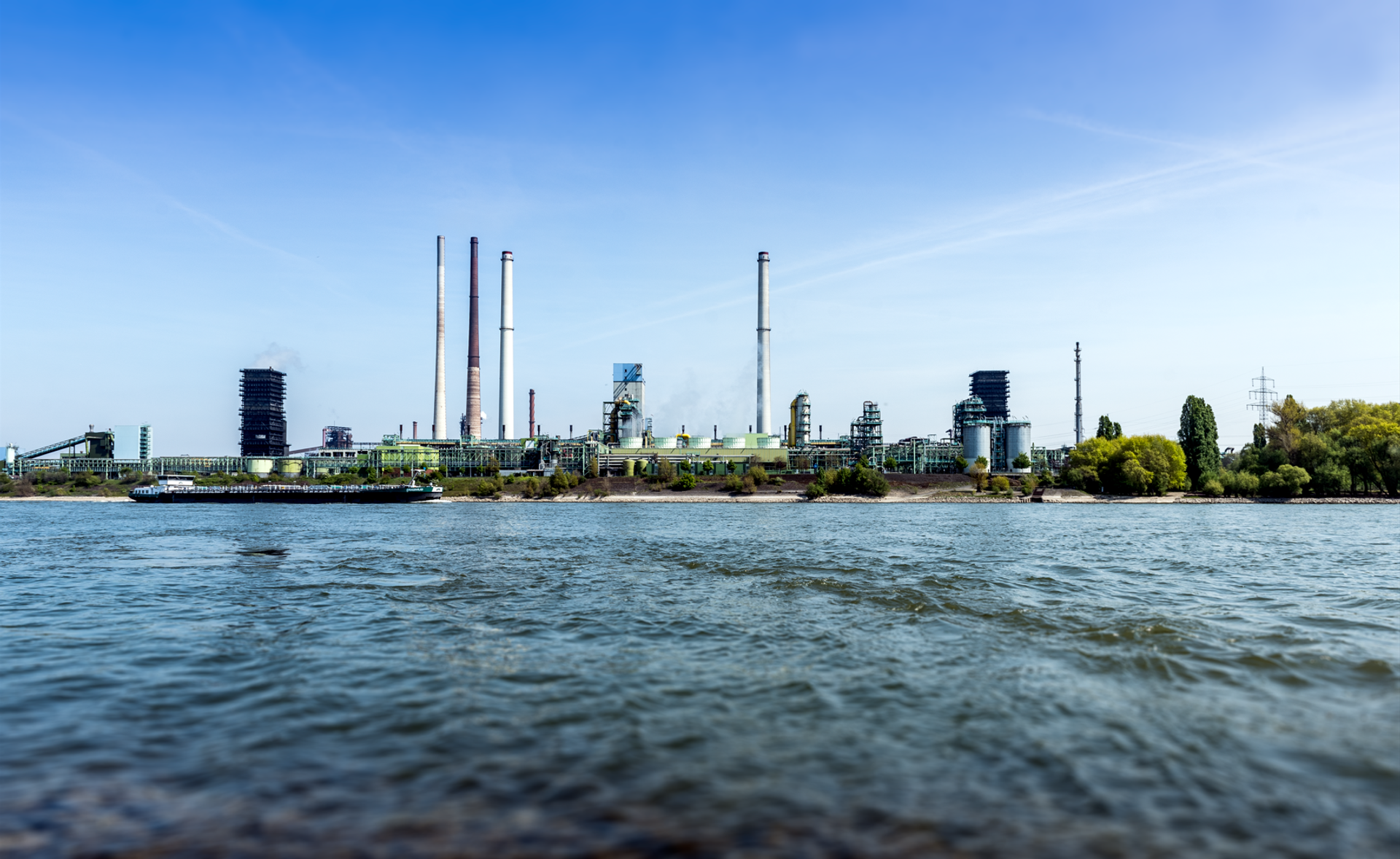 Extensive thyssenkrupp Steel site with industrial facilities and chimneys on the waterfront under a blue sky.