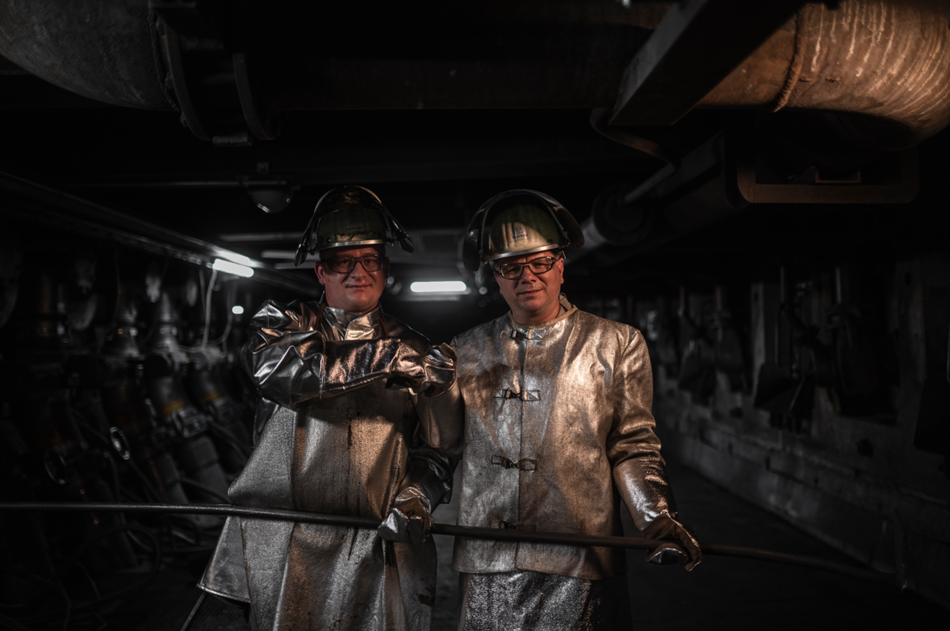 Zwei Mitarbeitende in silberner Schutzkleidung stehen in einem Industriebereich und blicken lächelnd in die Kamera.