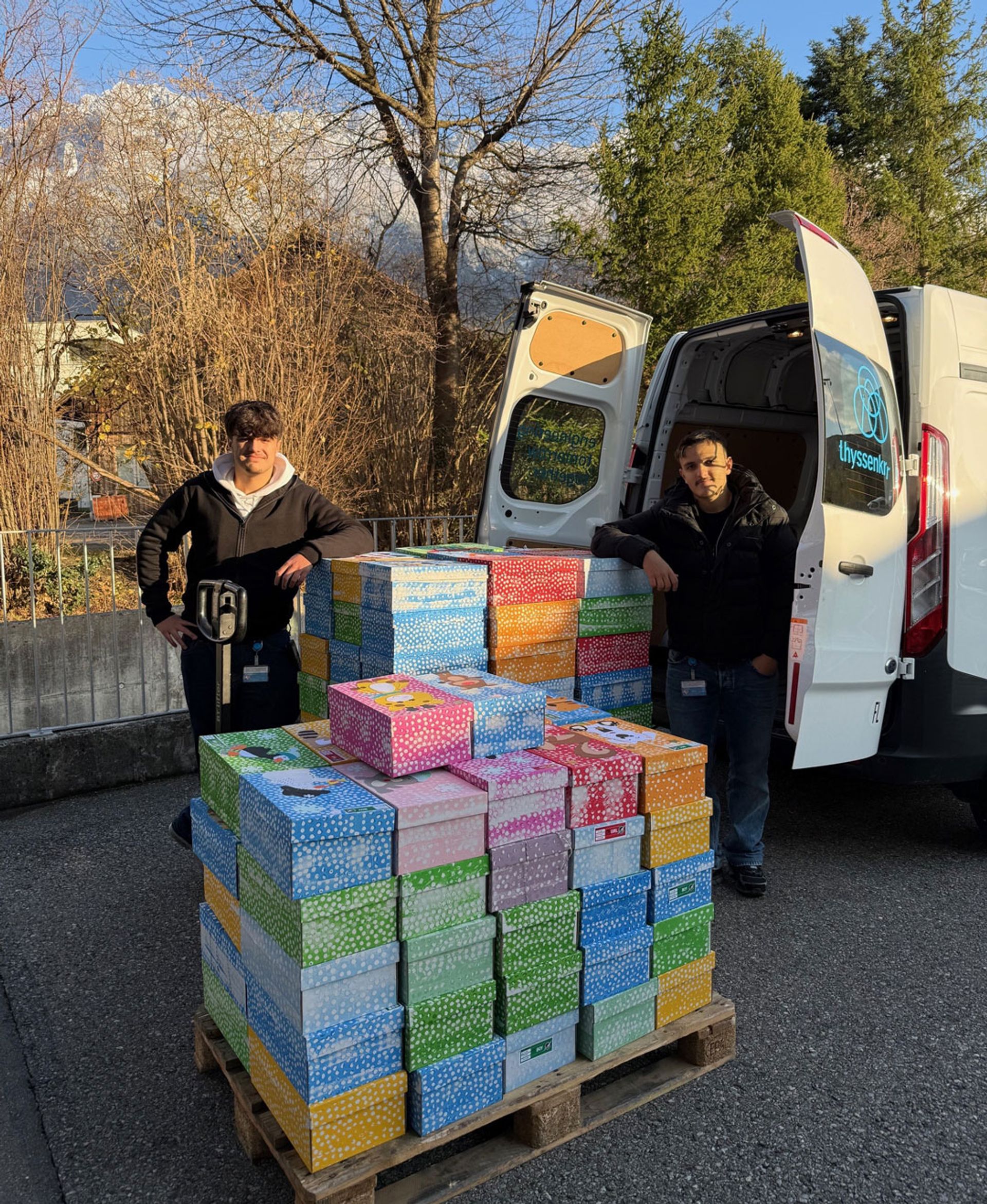 Zwei Auszubildende stehen an einem Firmenfahrzeug neben einem Stapel bunt verpackter Schuhkartons für „Weihnachten im Schuhkarton".