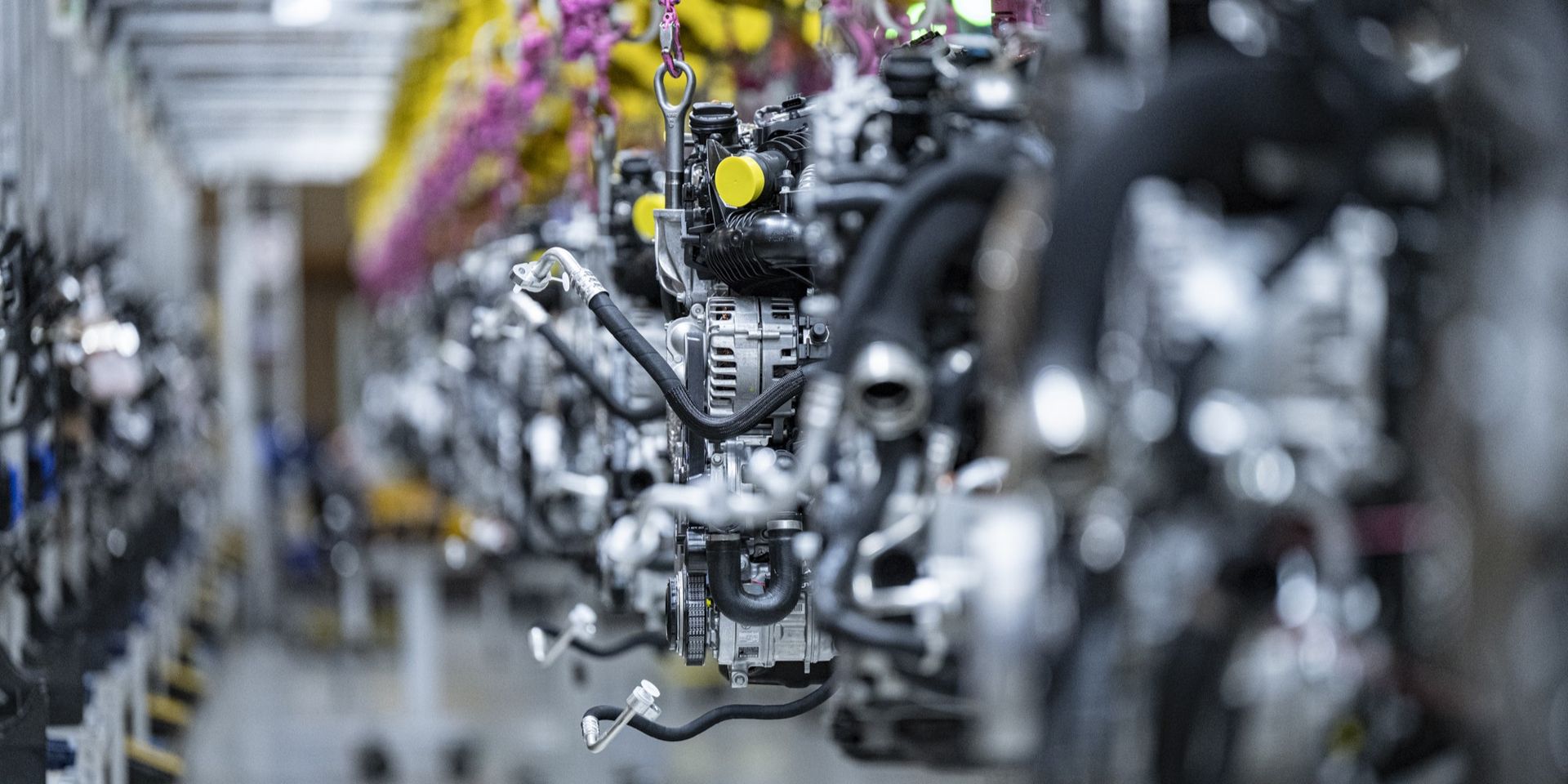 Mehrere Motoren hängen an einer Förderanlage in einer Produktionshalle; ein Motor in der Bildmitte ist scharf fokussiert, während die übrigen unscharf erscheinen.