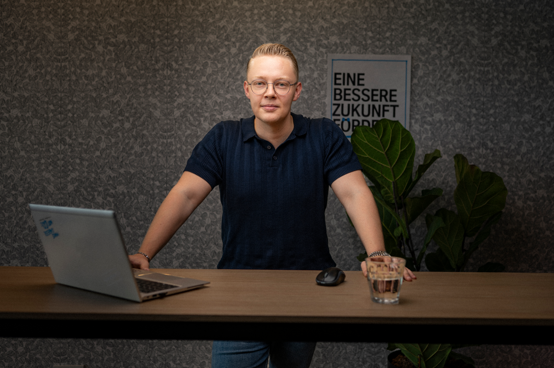 Ein Mitarbeiter steht an einem Schreibtisch, vor ihm ein Laptop und ein Glas Wasser. Er blickt in die Kamera, während im Hintergrund Pflanzen und ein Poster zu sehen sind.