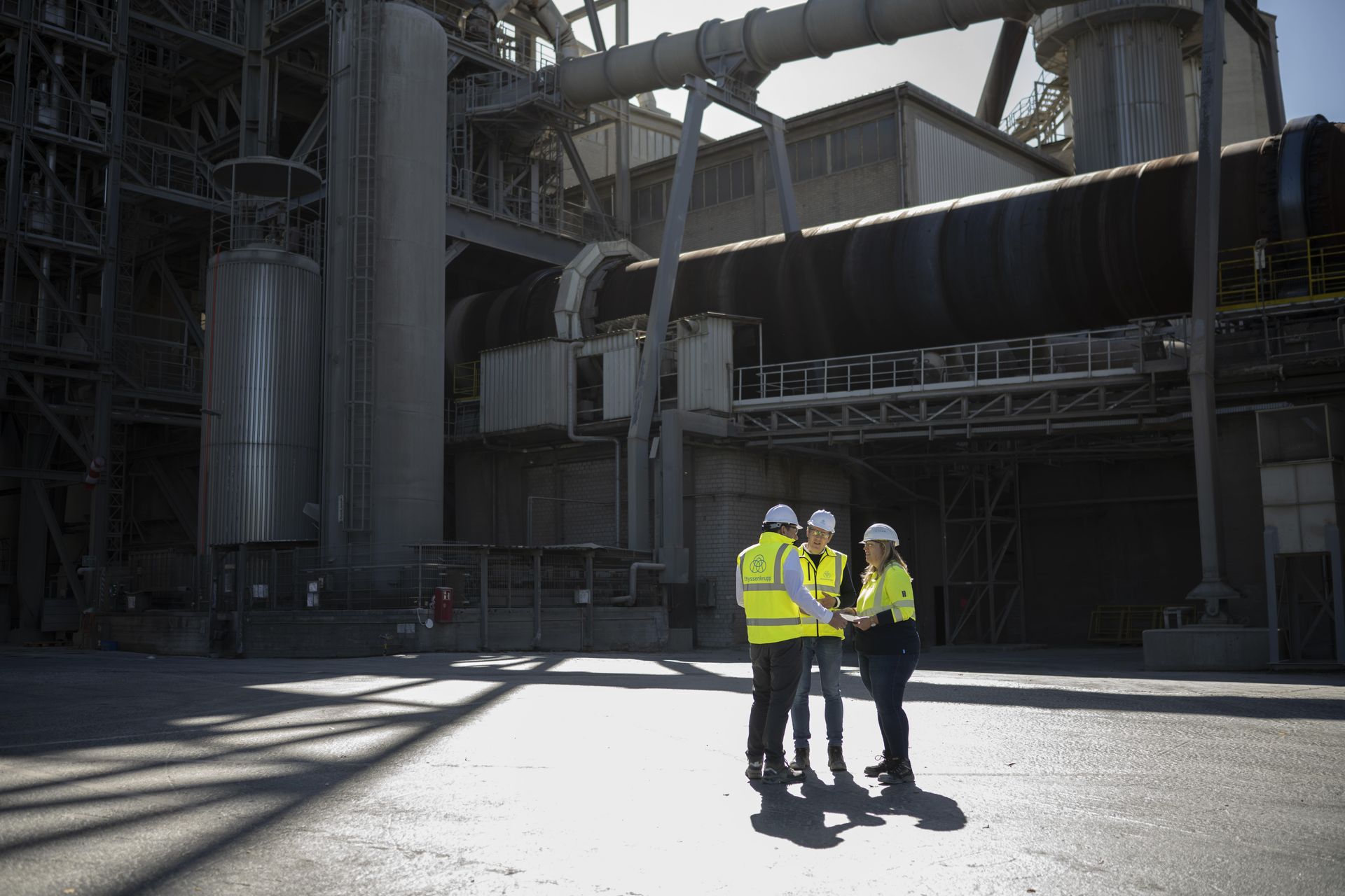 Drei thyssenkrupp Beschäftigte in Warnschutzkleidung stehen vor einer großtechnischen Industrieanlage.