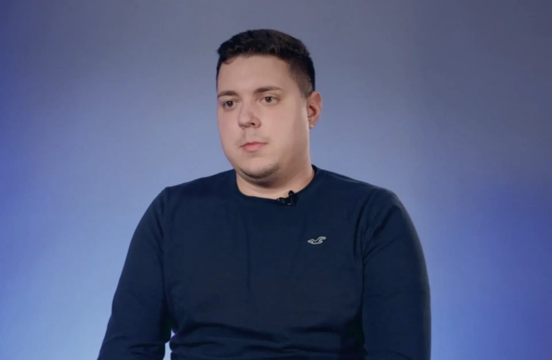 a visibly male-presenting employee in a dark navy jumper with a clip-on microphone, filmed against a blue studio background, sharing a personal statement about workplace health and safety