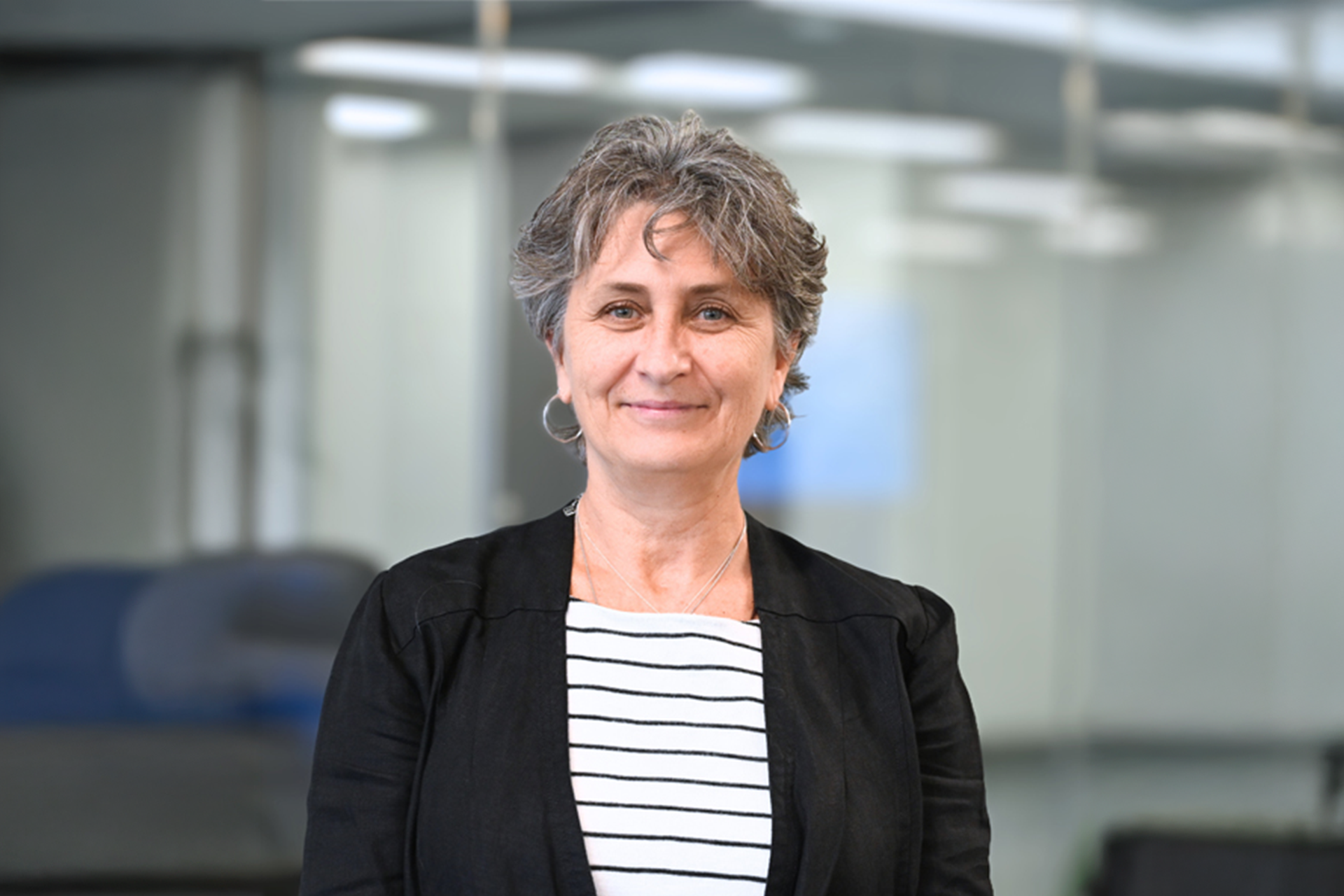Frau in schwarzem Blazer und gestreiftem Shirt steht vor moderner Glaswand im Büro.