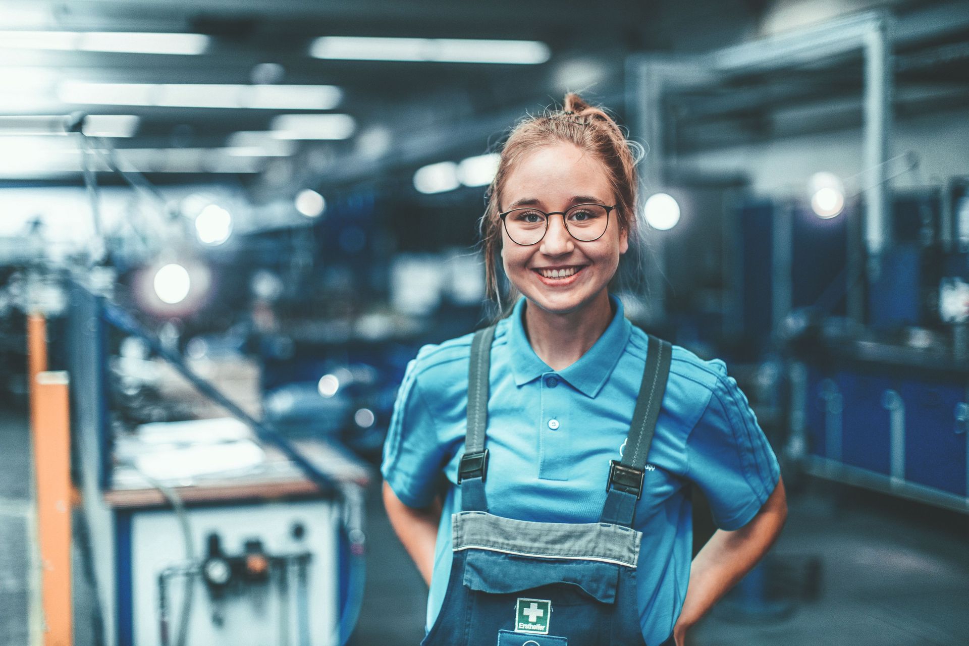 Eine lächelnde junge Frau in blauer Arbeitskleidung steht in einer modernen Werkstatt. Im Hintergrund sind unscharf Maschinen und Arbeitsbereiche zu sehen.