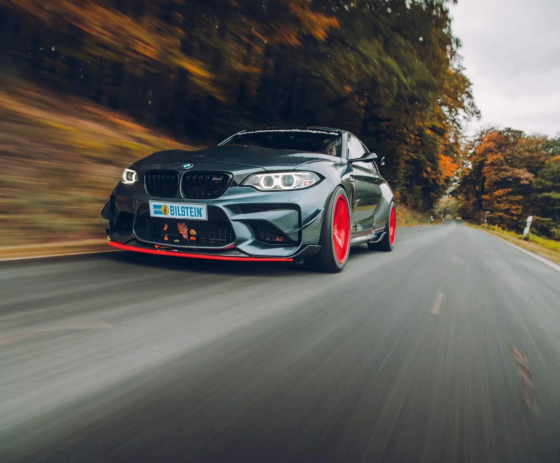 Ein dunkelgraues sportliches Coupé von BMW mit roten Felgen fährt auf einer Landstraße durch eine herbstliche Landschaft.