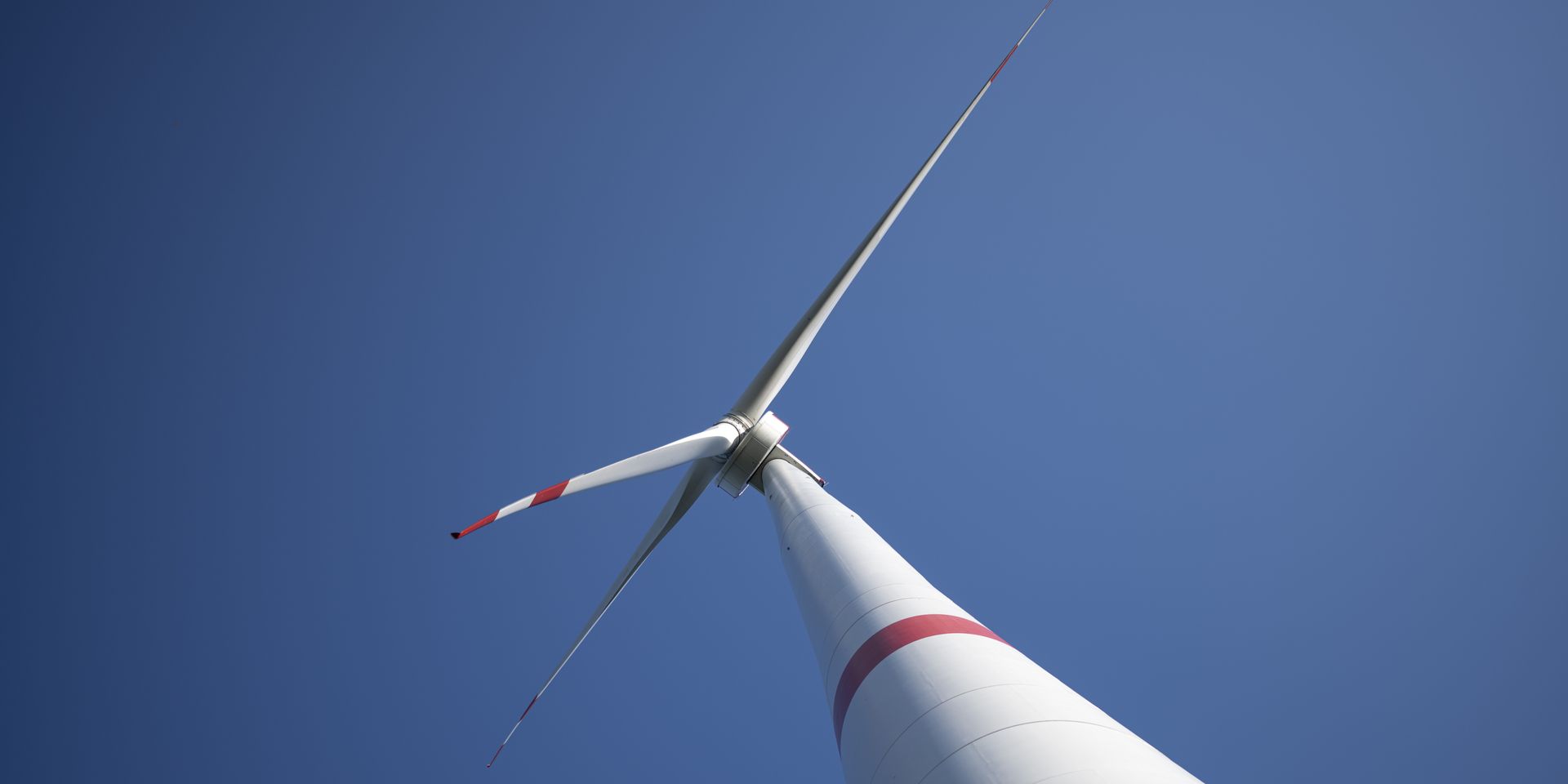 Blick entlang des Turms auf die Gondel und die Rotorblätter einer Windkraftanlage vor blauem Himmel.