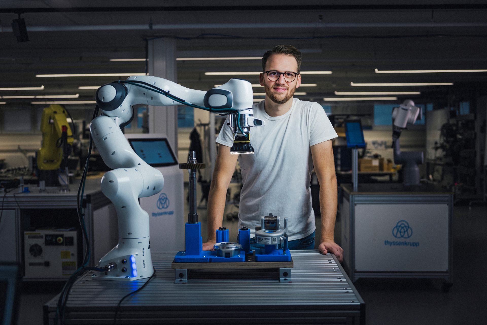 Mann steht in einem Techniklabor neben einem weißen Roboterarm, auf einem Tisch liegen Werkstücke und Messgeräte.