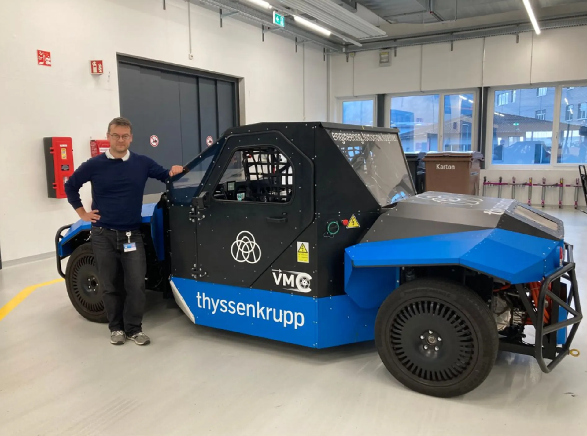 Männlich gelesene Person steht in einer Werkhalle neben einem blau-schwarzen Fahrzeugprototyp mit thyssenkrupp Logo.