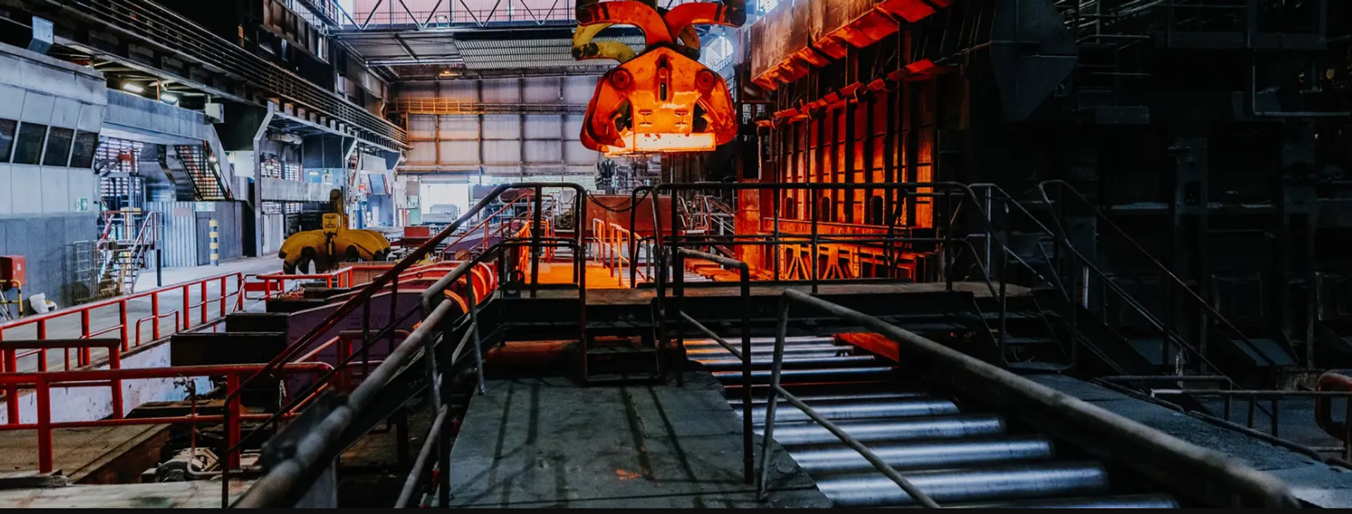 Industriehalle eines Stahlwerks mit großen Produktionsanlagen, Förderrollen und einem orange leuchtenden Greifer über dem Arbeitsbereich.