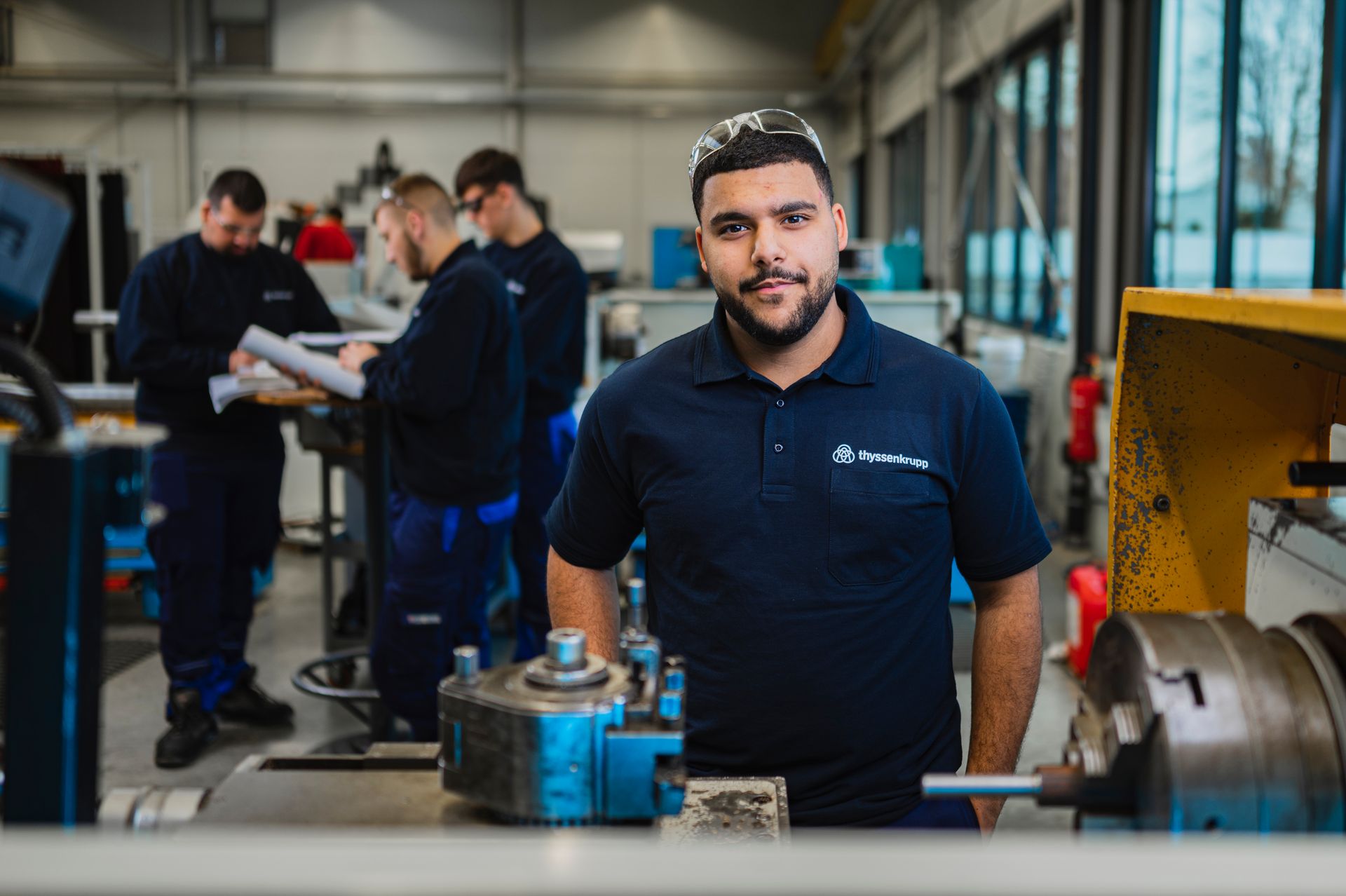 Ein junger Mann in Arbeitskleidung steht vor einer Metallmaschine, während Kollegen im Hintergrund an technischen Unterlagen arbeiten.