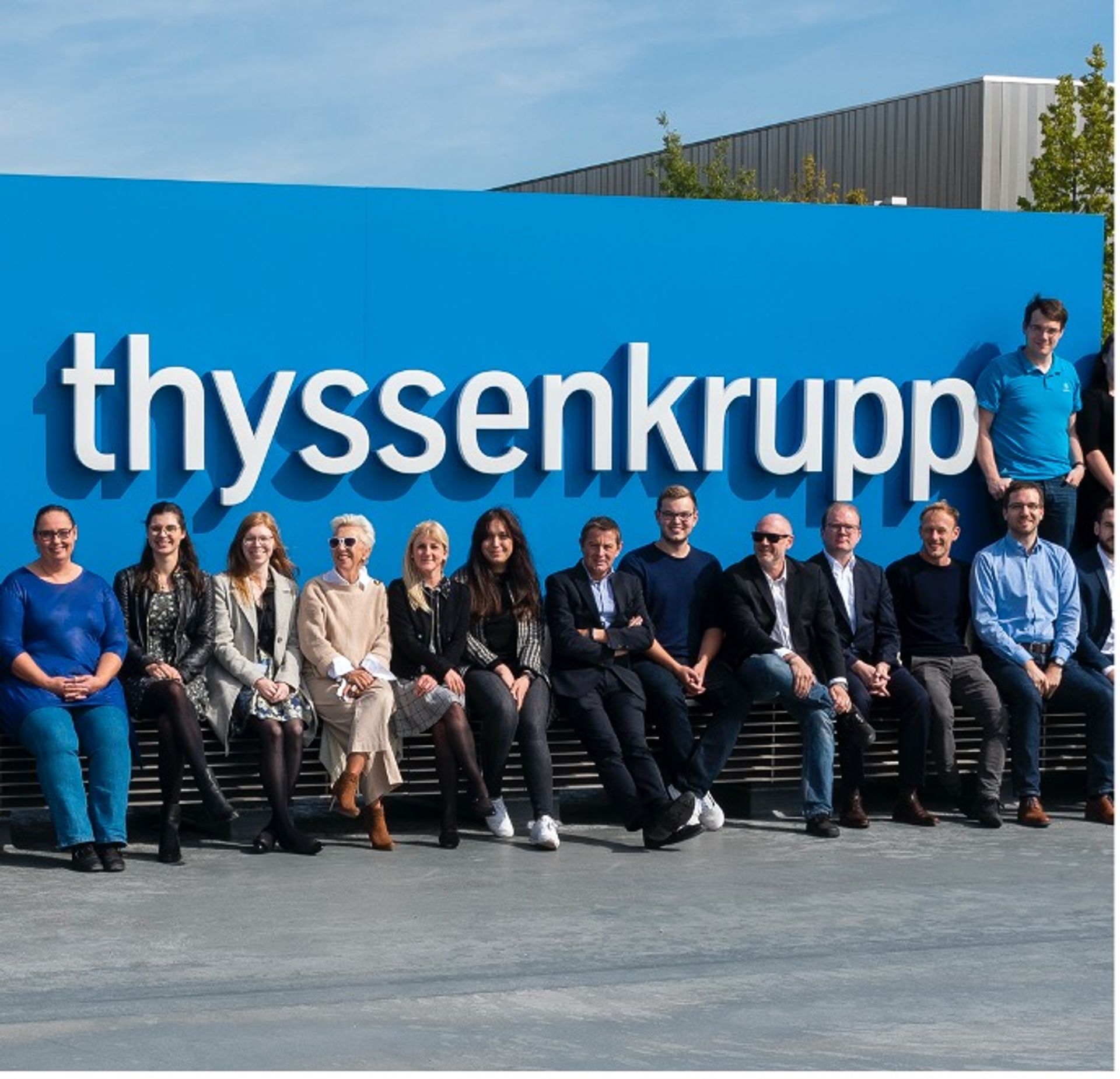 Mitarbeitende von thyssenkrupp vor einem großen thyssenkrupp Logo. 