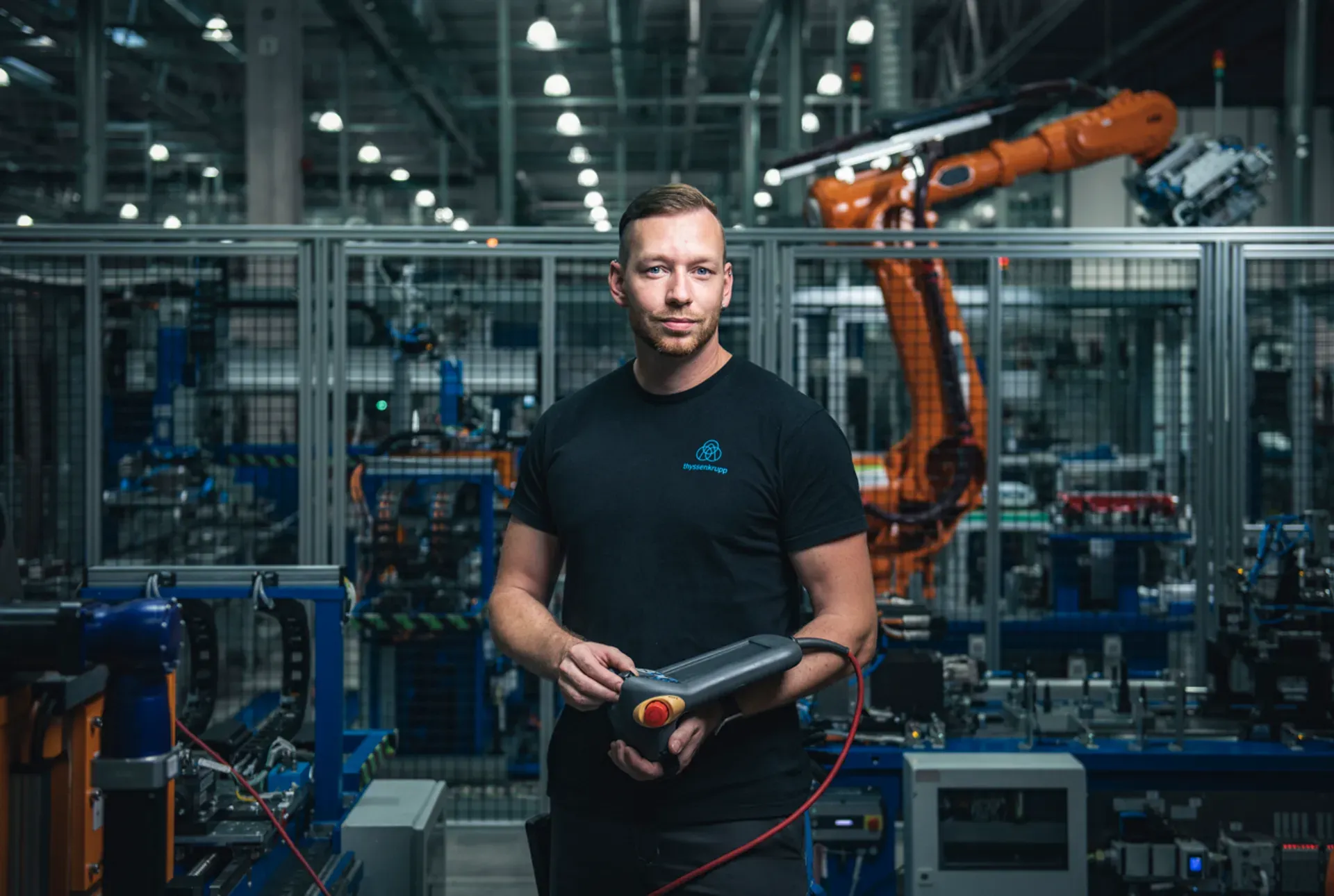 A person is holding a control unit in an industrial environment with machines in the background.