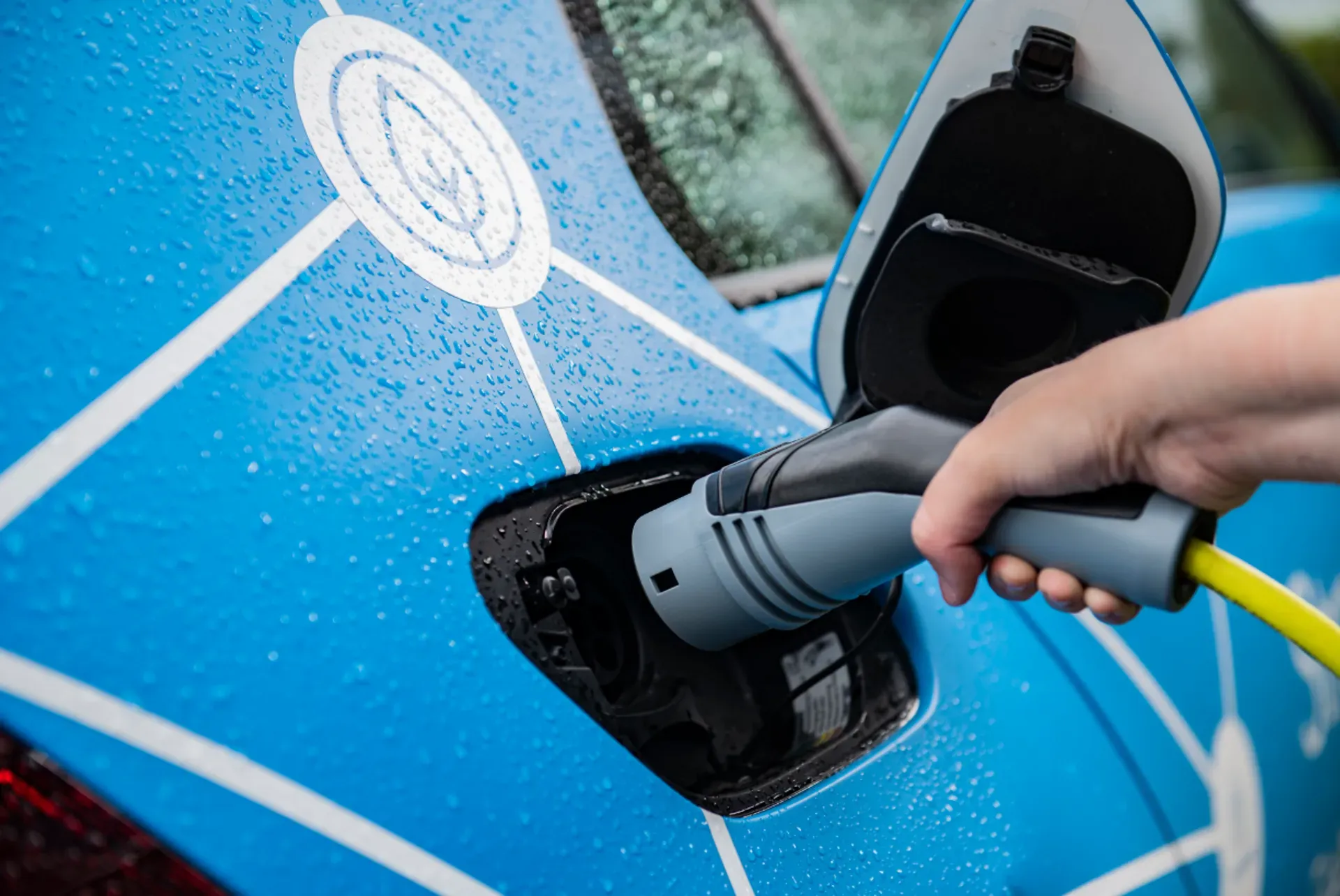Eine Hand steckt einen Ladestecker in den Ladeanschluss eines blauen Elektrofahrzeugs; auf der Karosserie sind Regentropfen zu sehen.