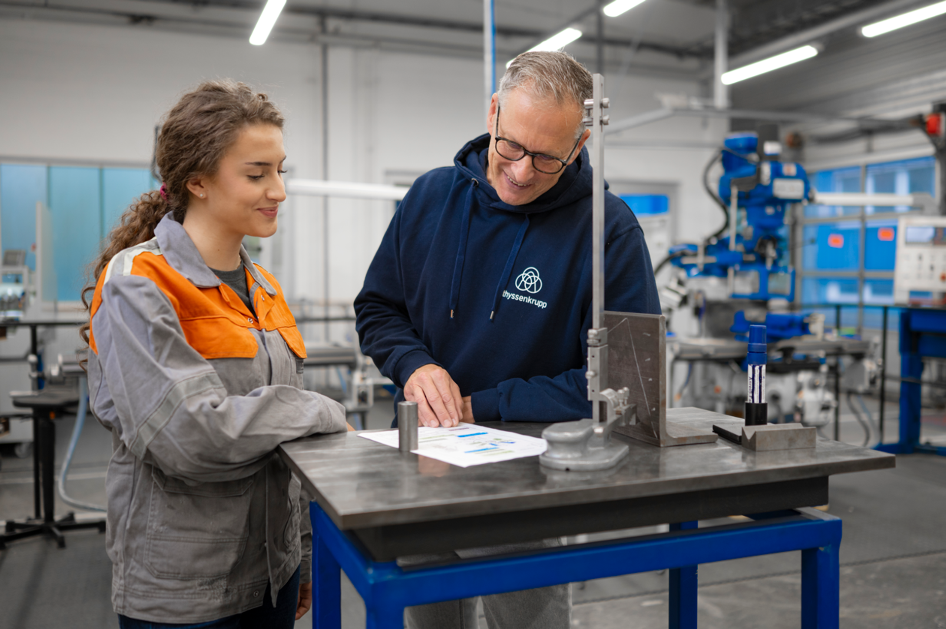A thyssenkrupp instructor discusses technical drawings with an apprentice at a workbench in a modern industrial workshop.