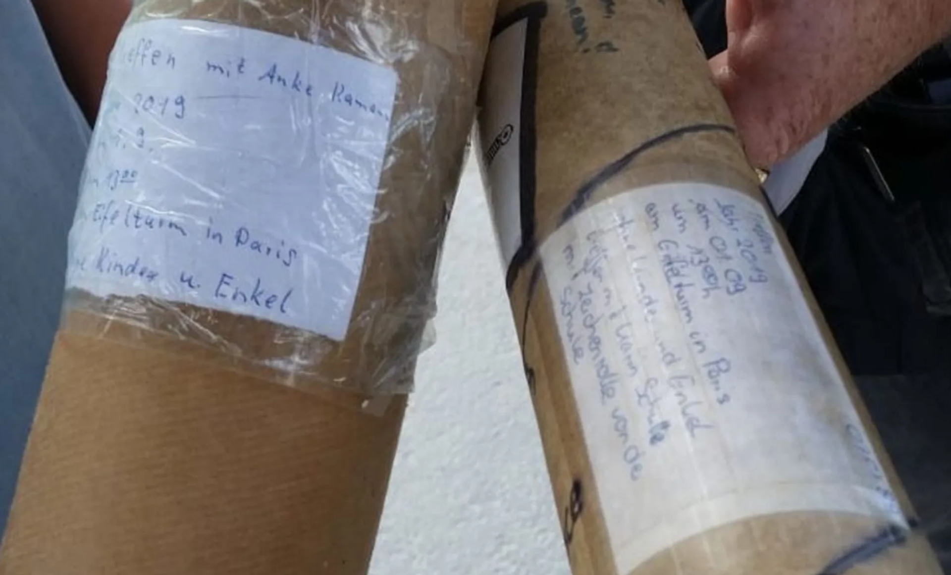 Close-up of two long cardboard packages with handwritten labels, held by people.