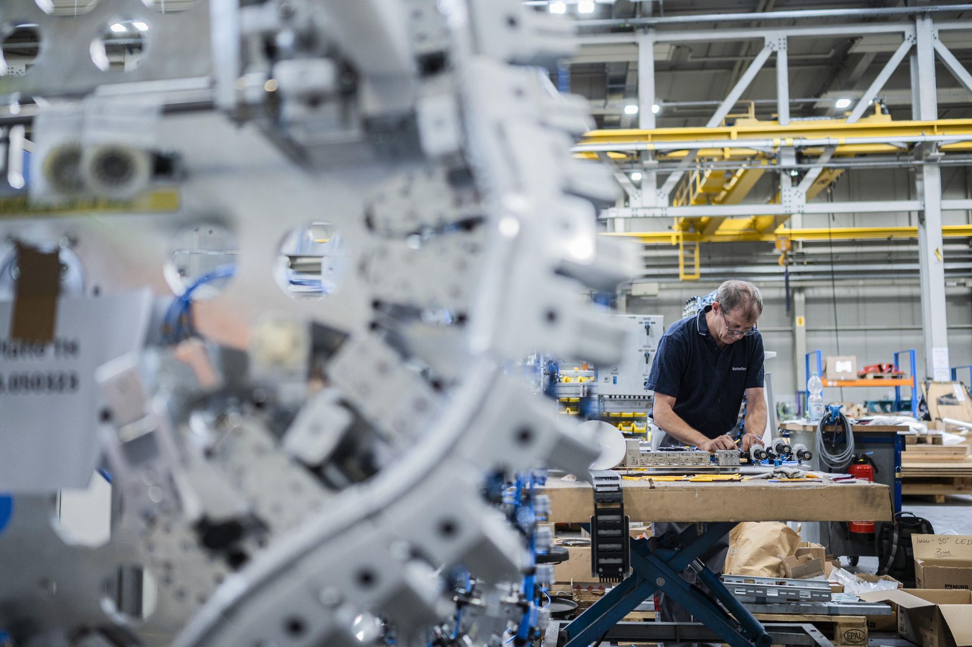 Ein Mitarbeiter arbeitet an einem Montagetisch in einer industriellen Fertigungshalle für Automobilkomponenten; im Vordergrund ist eine große Maschinenanlage unscharf zu sehen.