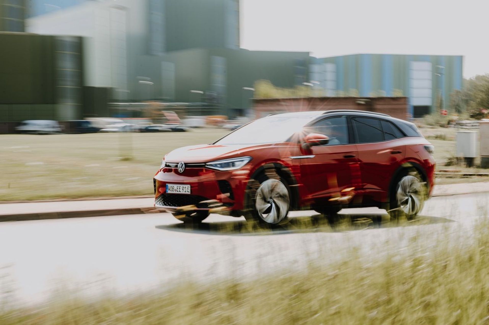 A red electric car drives down a street, passing an industrial building.
