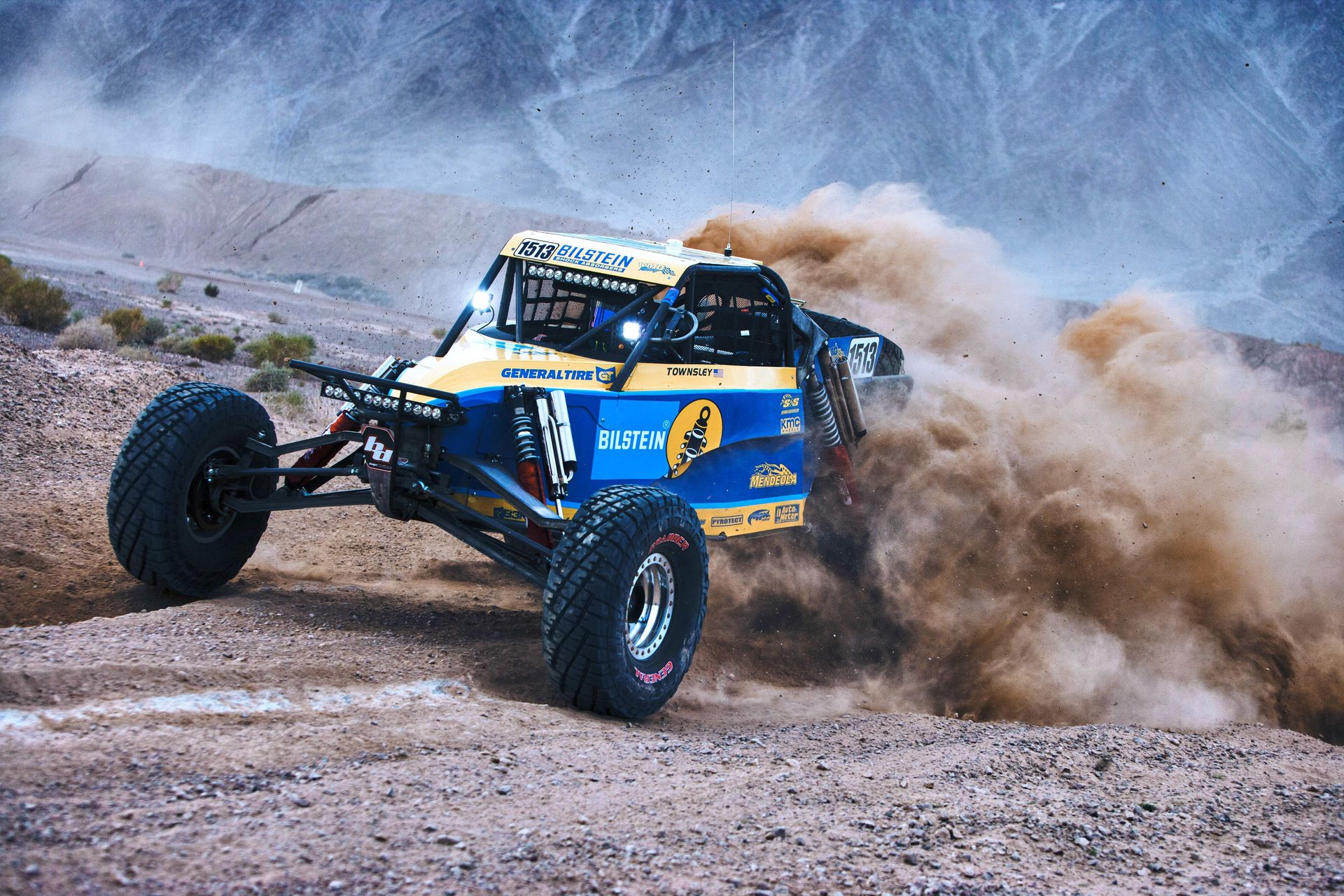 Ein Off-Road-Fahrzeug von BILSTEIN fährt mit hoher Geschwindigkeit über eine steinige Wüstenstrecke und wirbelt dabei eine große Staubwolke auf.