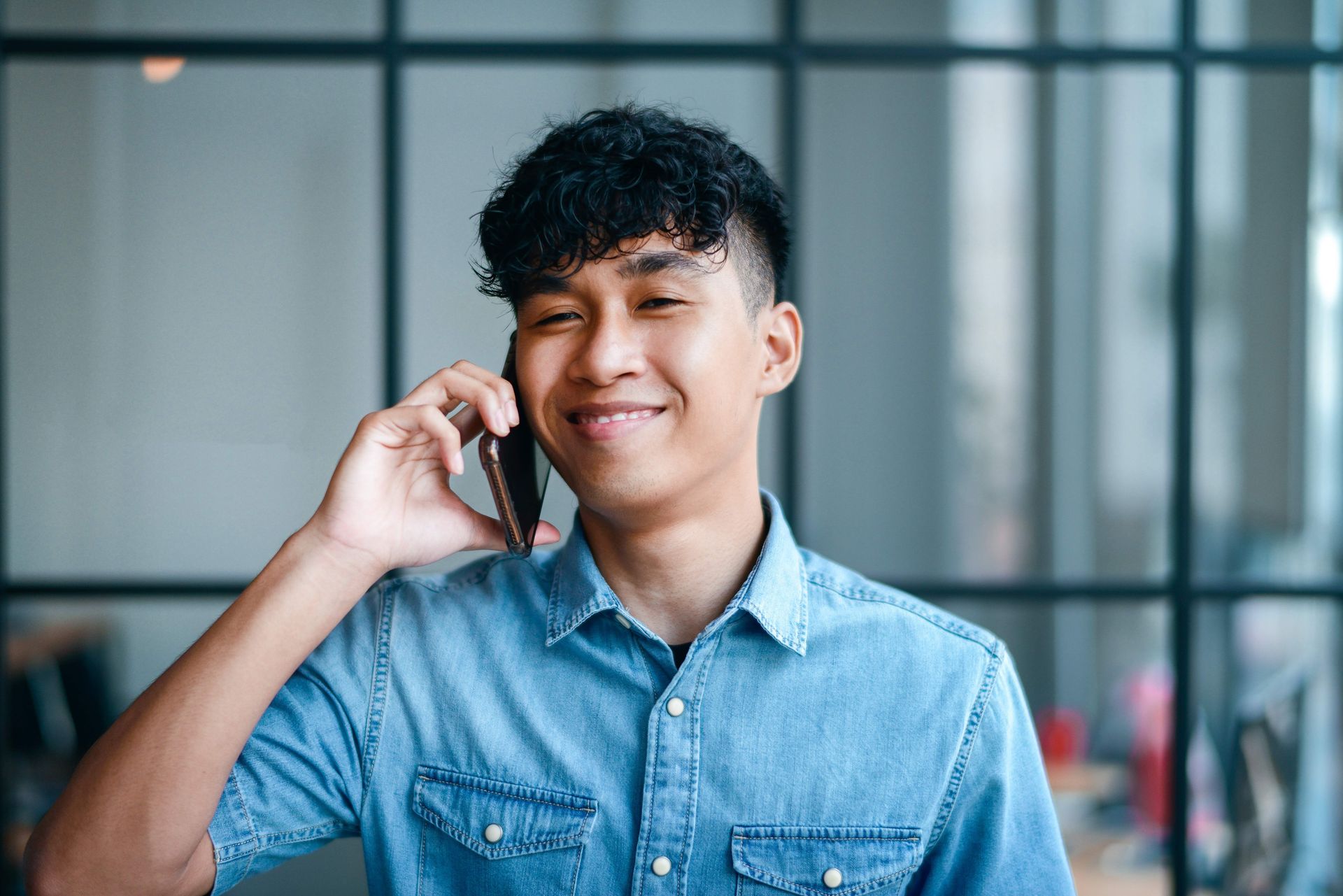 Ein junger Mann steht in einem modernen Innenraum und führt ein Telefonat. Er trägt ein helles Jeanshemd und lächelt freundlich, während er das Smartphone an seinem Ohr hält. Hinter ihm sind große Fenster mit einem schlichten Metallrahmen zu sehen, die der Szene ein helles, offenes Ambiente verleihen.