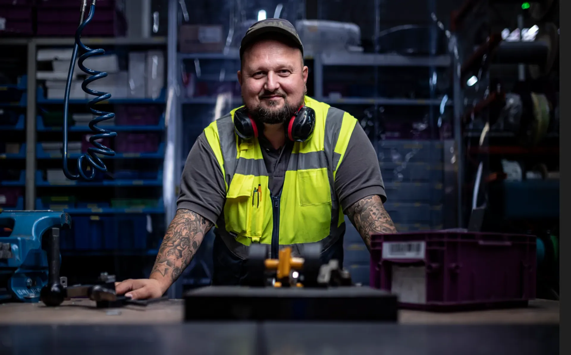 a visibly male-presenting employee with tattoos, wearing a high-visibility vest and hearing protection around the neck, smiling at the camera while leaning on a workbench in an industrial warehouse