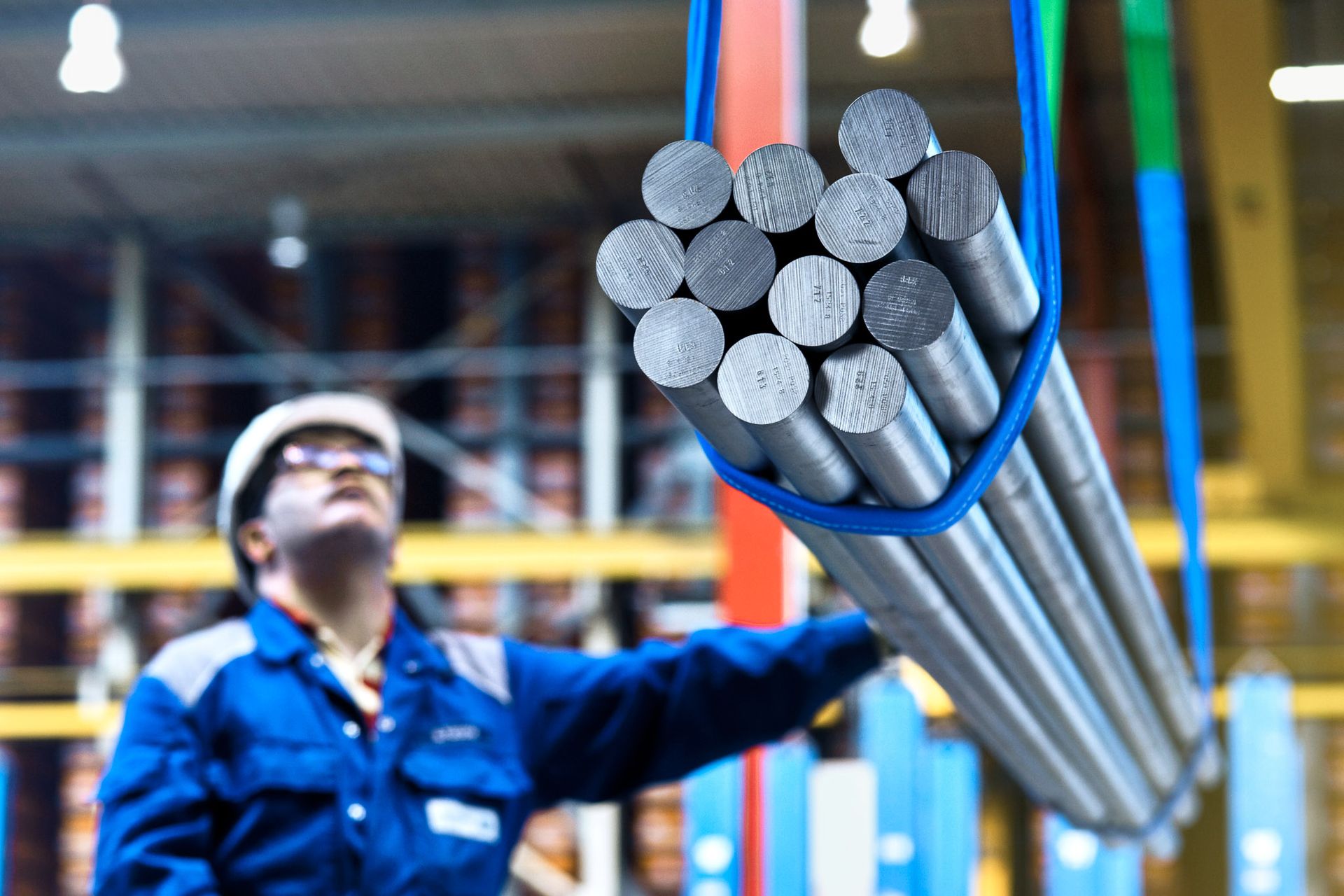 Mehrere Metallstangen hängen an blauen Gurten in einer Lagerhalle.