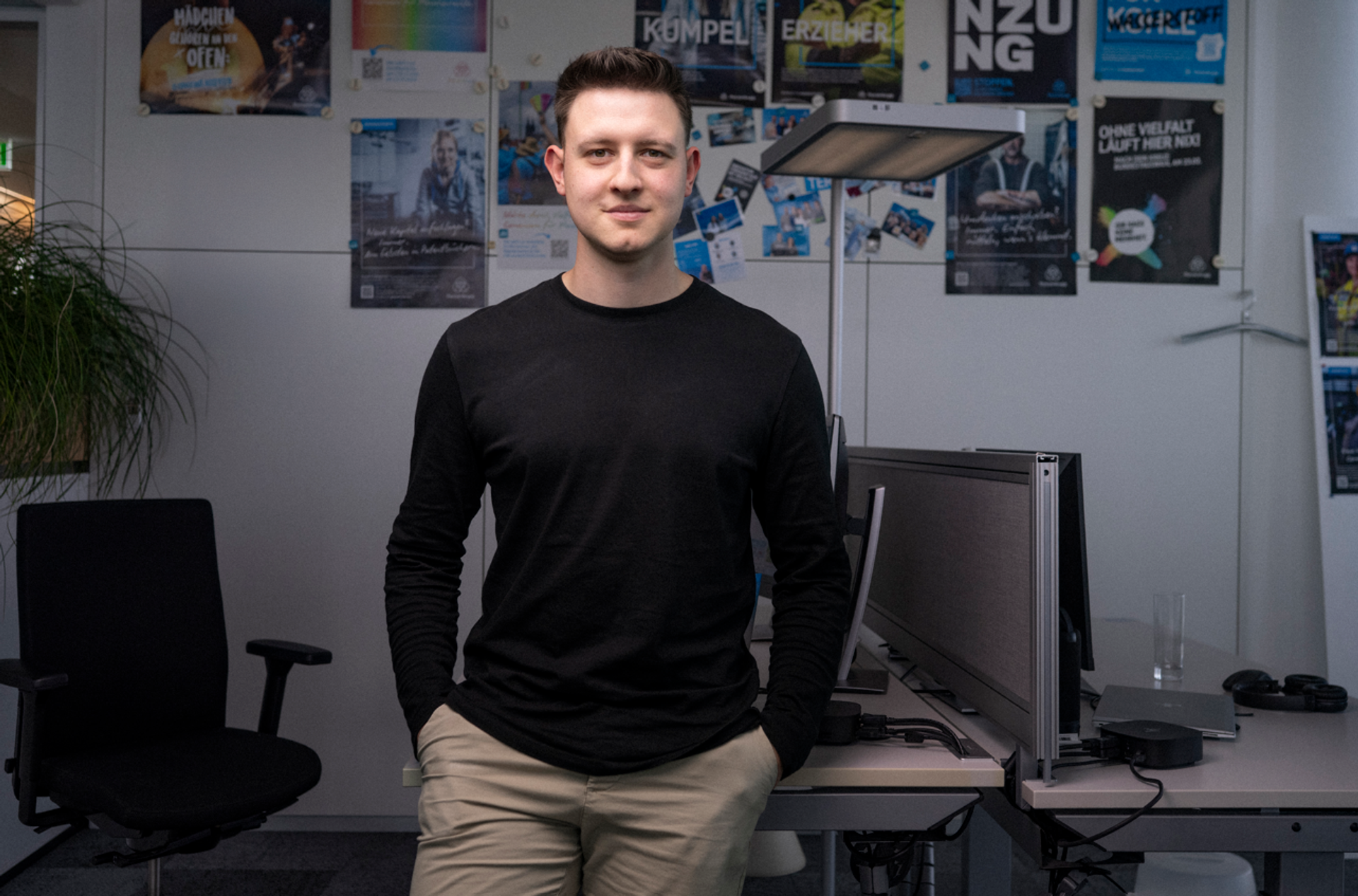 Junger Mann steht in einem Büro mit verschiedenen Plakaten an der Wand und lächelt in die Kamera.
