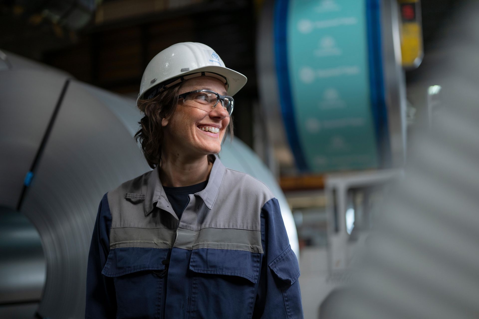 An employee wearing a hard hat is standing in a production hall in front of a large coiled steel roll.
