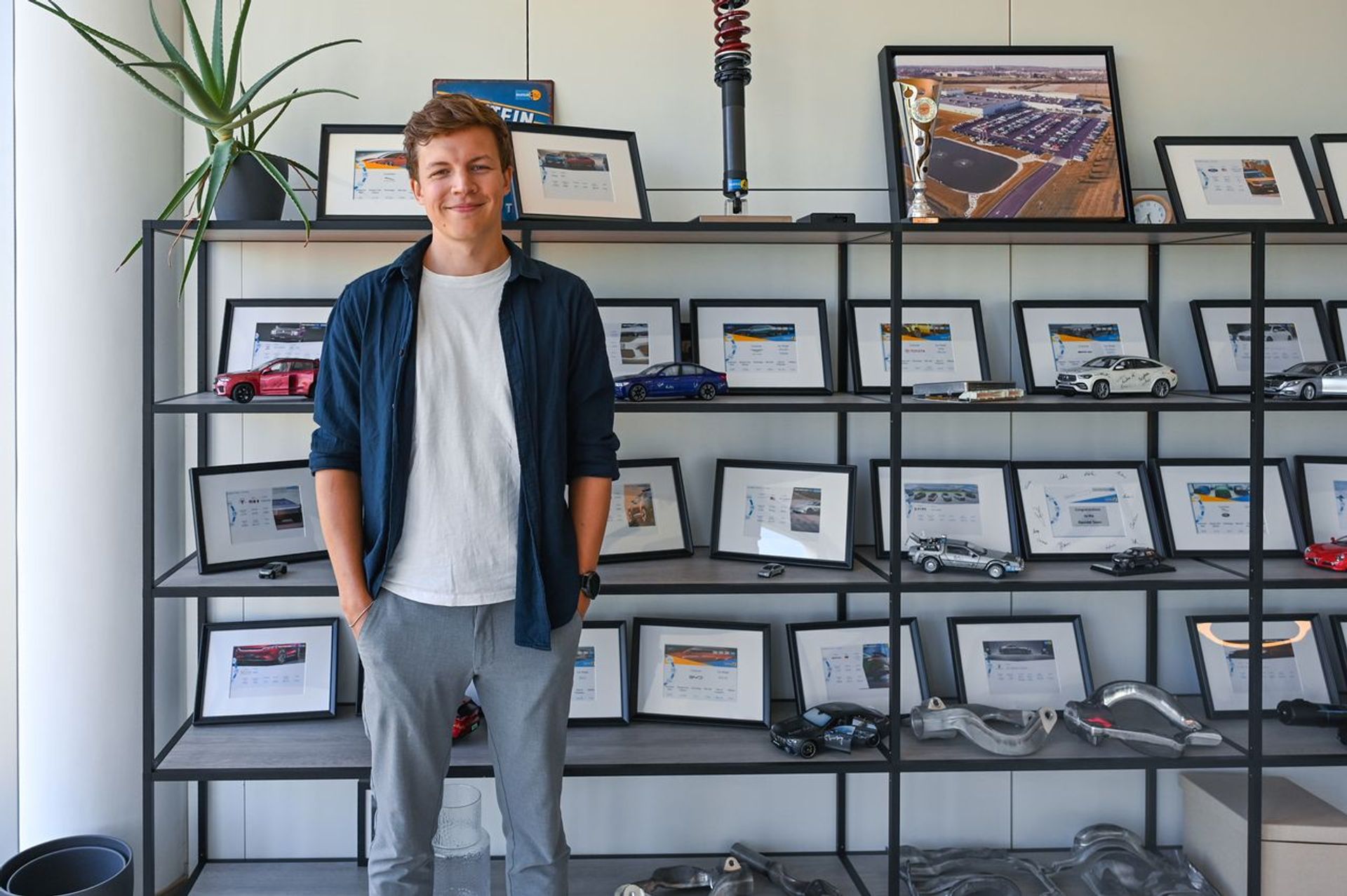 Felix Ewering von thyssenkrupp Bilstein steht vor einem Regal mit Fahrzeugmodellen, technischen Bauteilen und gerahmten Projektmotiven.