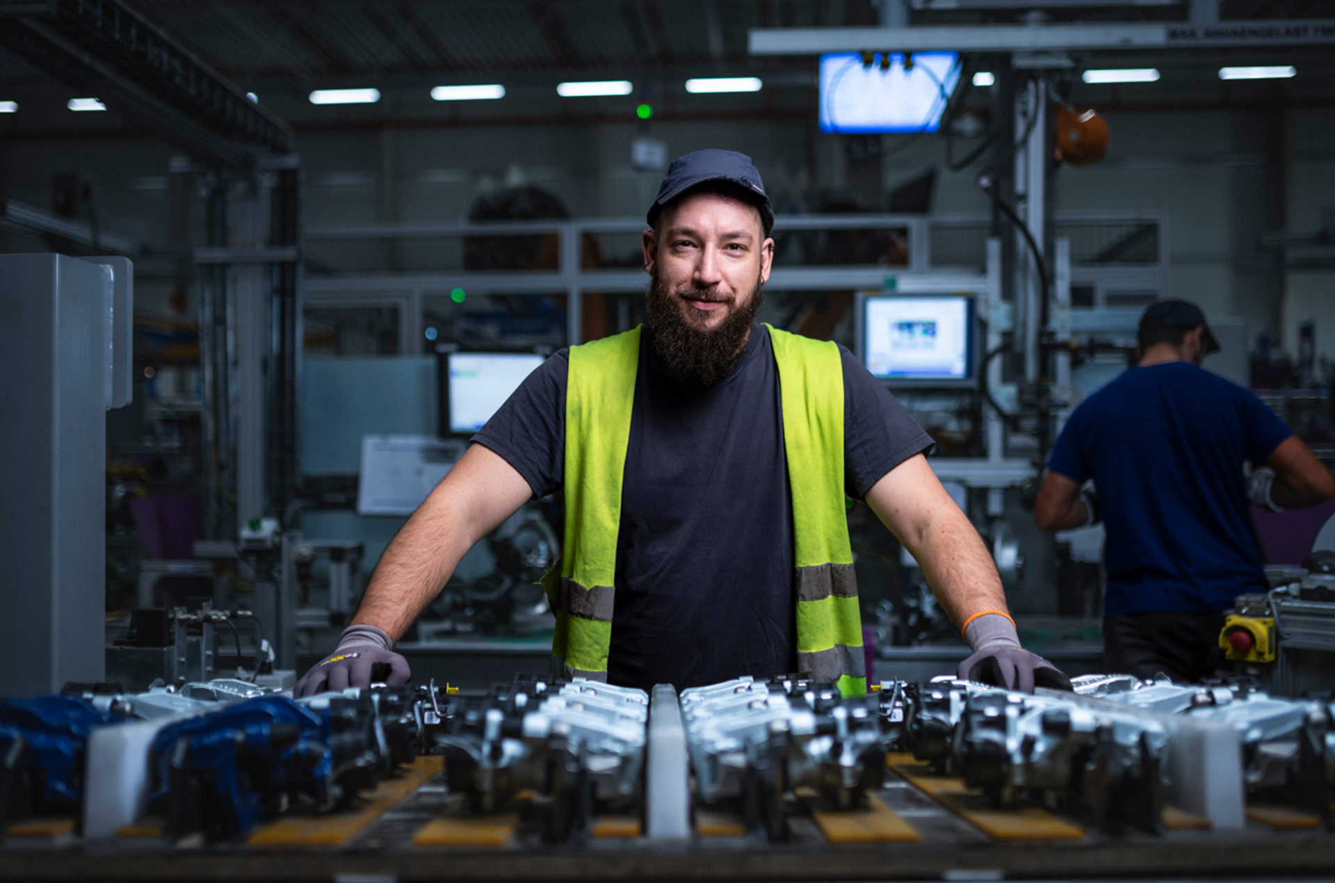 Mann steht an einer Produktionslinie in einer Fabrikhalle mit Maschinen und Bauteilen.