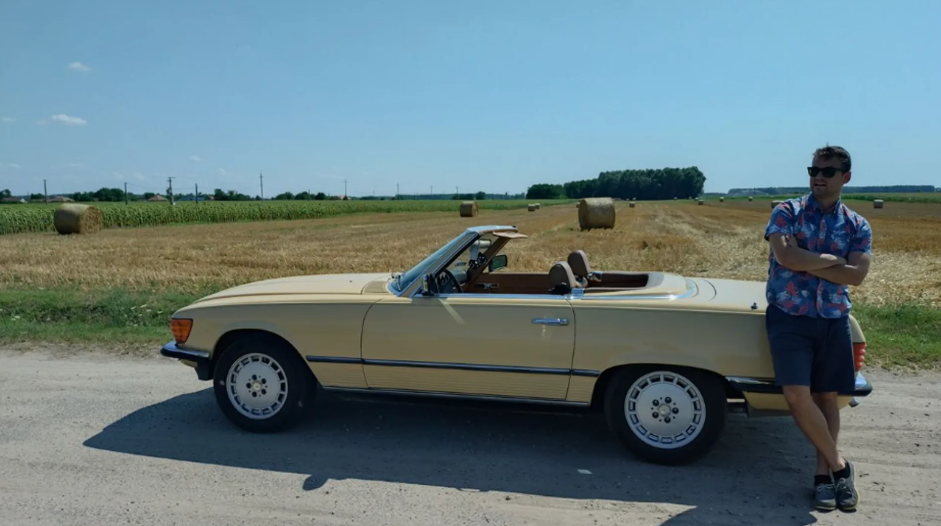 Männlich gelesene Person mit Sonnenbrille lehnt mit verschränkten Armen an einem beigefarbenen Cabriolet vor einem Feld mit Heuballen.