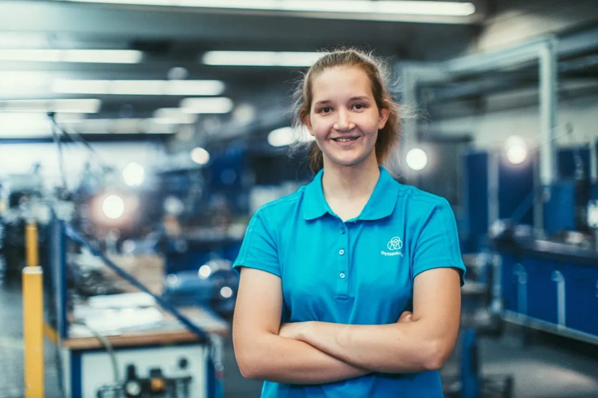 Junge Frau in blauer Arbeitskleidung steht mit verschränkten Armen in einer Werkhalle mit Maschinen im Hintergrund.