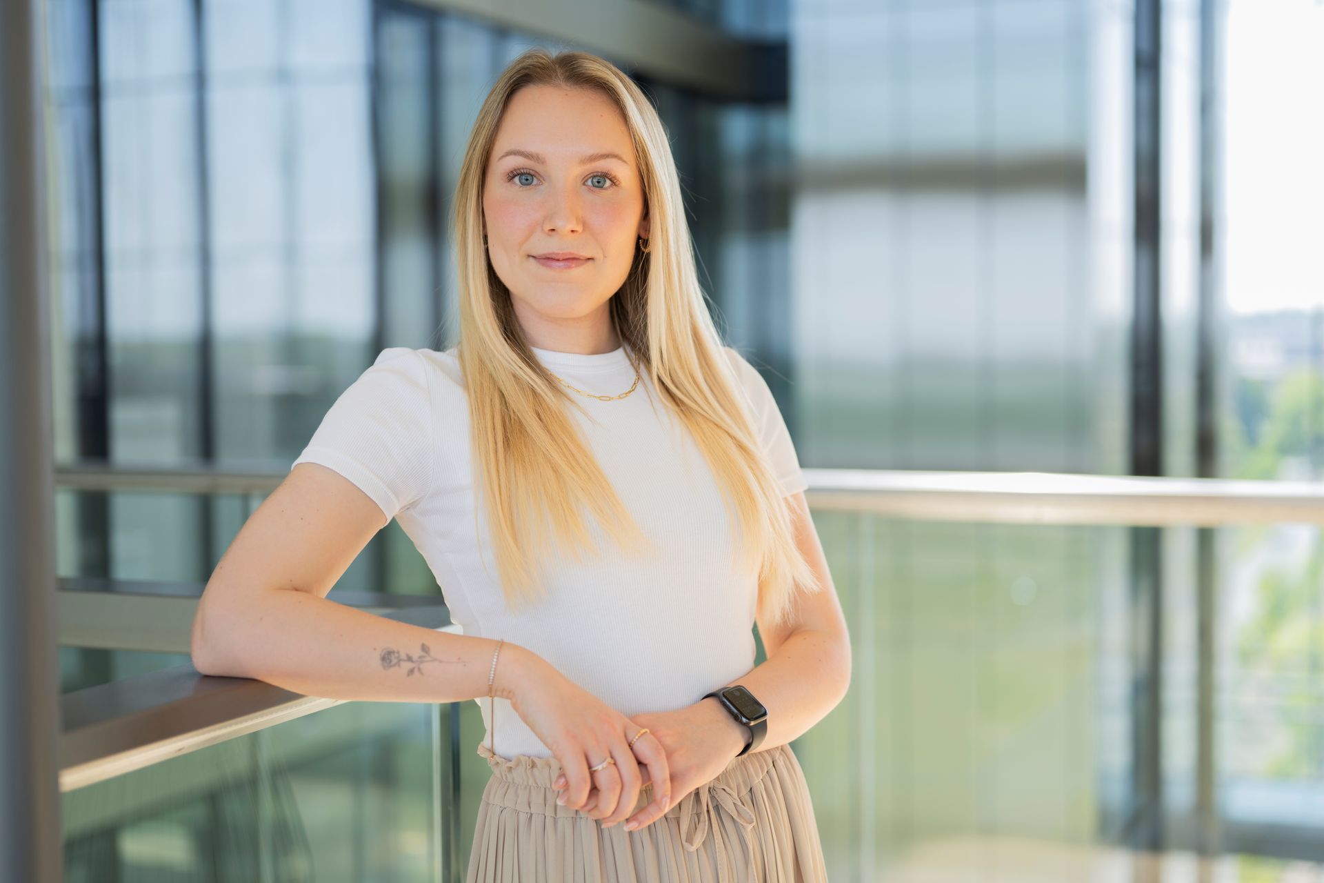 Frau steht vor einer Glasfassade in einem lichtdurchfluteten Bürogebäude, trägt helles Oberteil und Armbanduhr.