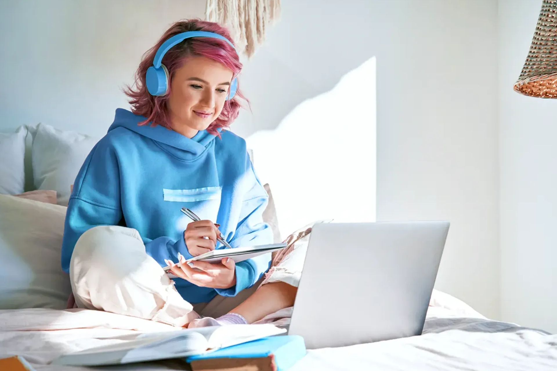 Person sitzt auf einem Bett mit Laptop und Notizblock, trägt Kopfhörer und arbeitet in einer hellen, gemütlichen Umgebung.