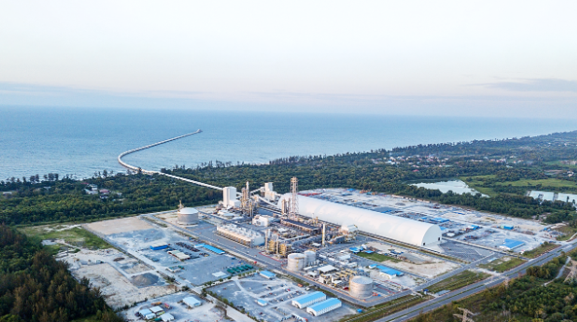 Ein rechteckig angelegtes Industriegebiet umgeben von grün bewachsenen Flächen, aus der Vogelperspektive, das Meer im Hintergrund