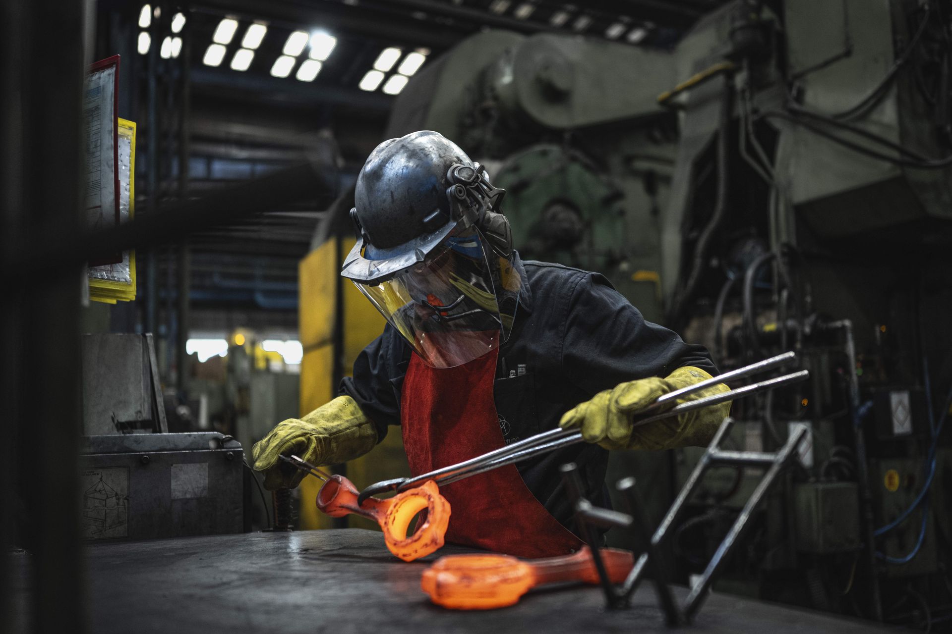 A thyssenkrupp employee wearing a hard hat, face shield, apron, and gloves is working on red-hot automotive components with long pliers at an industrial workstation.