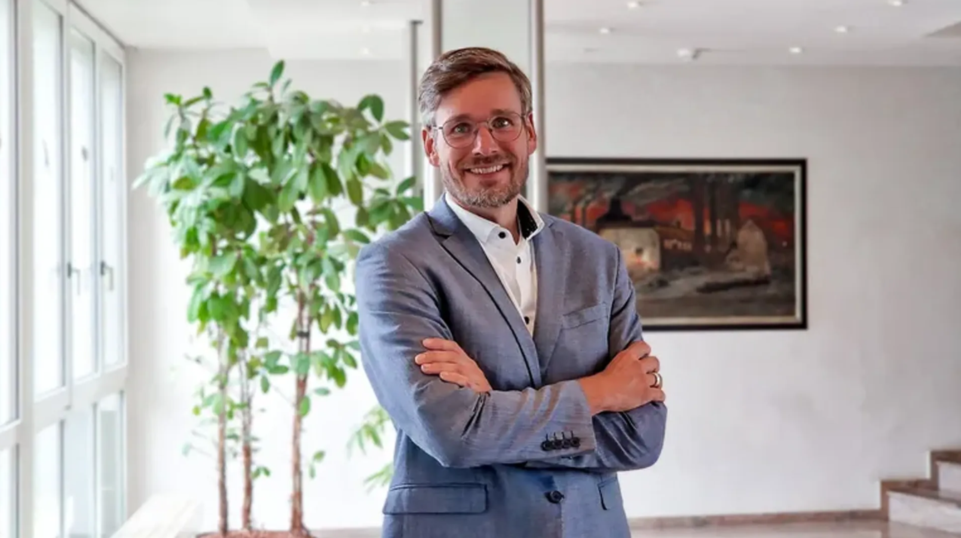 Männlich gelesene Person mit Brille und Anzug steht mit verschränkten Armen in einem hellen Bürogebäude; im Hintergrund sind eine große Pflanze und ein gerahmtes Bild zu sehen.