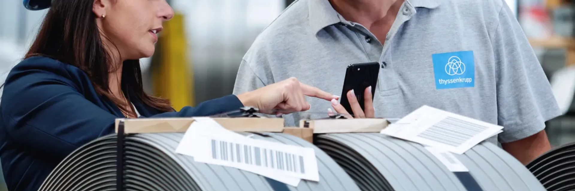 Two thyssenkrupp employees discussing information on a smartphone next to coiled metal strips in a production environment.