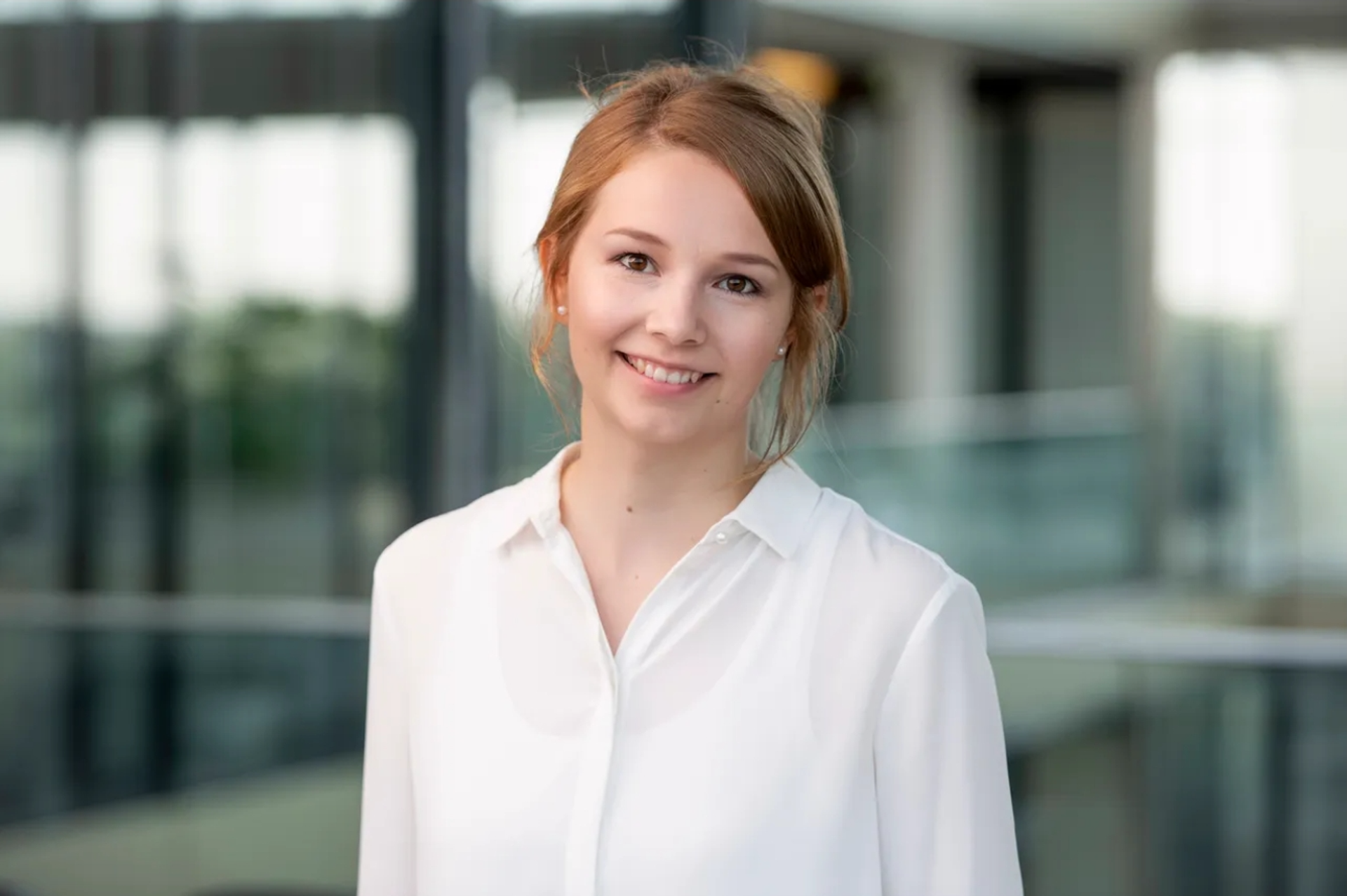 Lächelnde Frau in weißem Hemd steht in einem modernen Bürogebäude.
