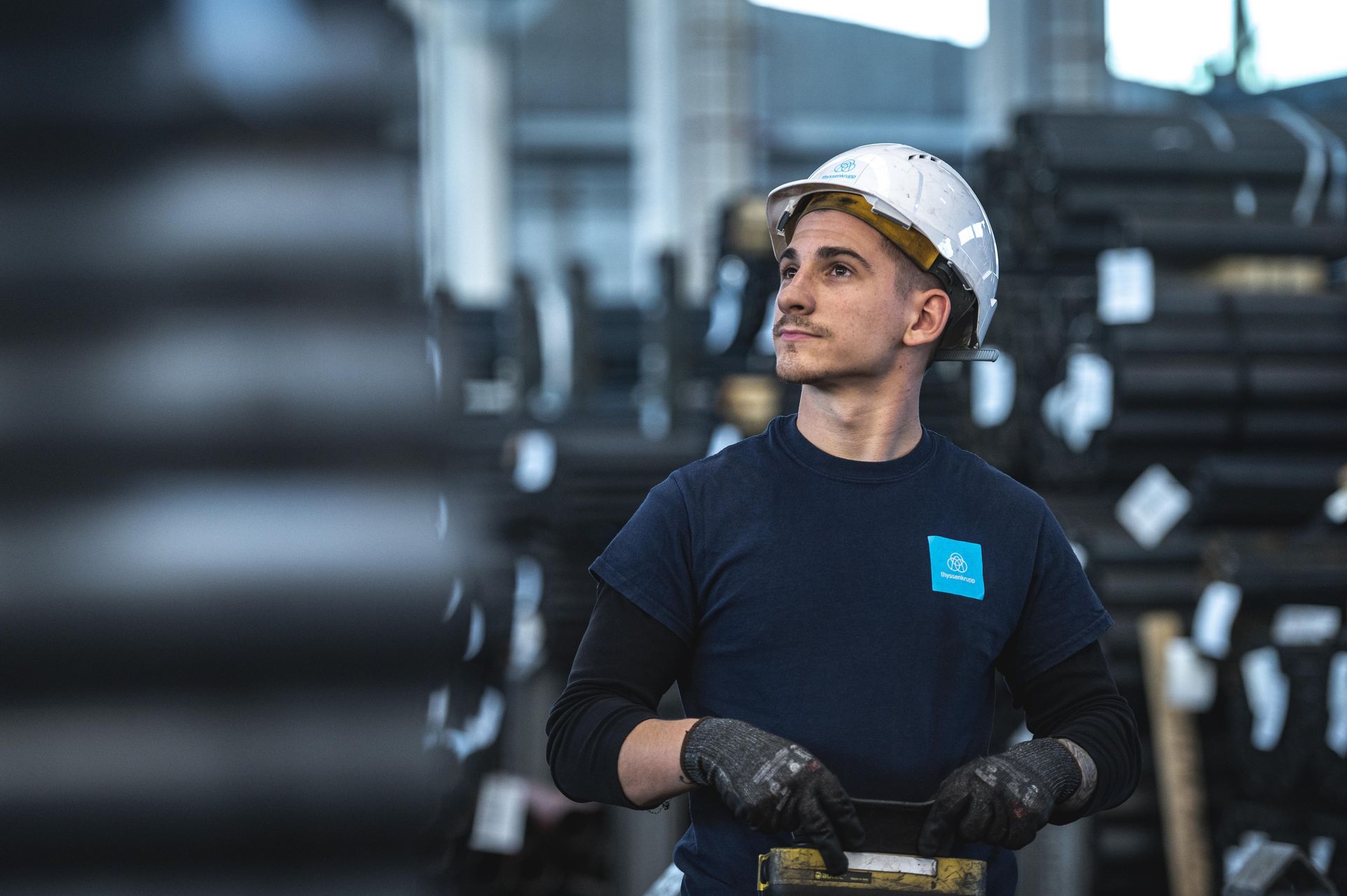 thyssenkrupp Mitarbeiter mit Schutzkleidung vor gelagerten Metallrollen in einer Produktionshalle.