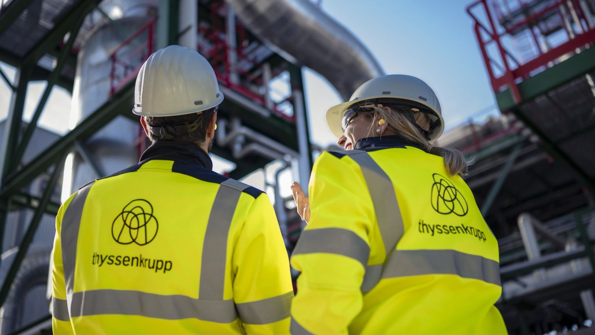 Zwei thyssenkrupp Mitarbeitende vor einer Industrieanlage mit Rohrleitungssystemen.