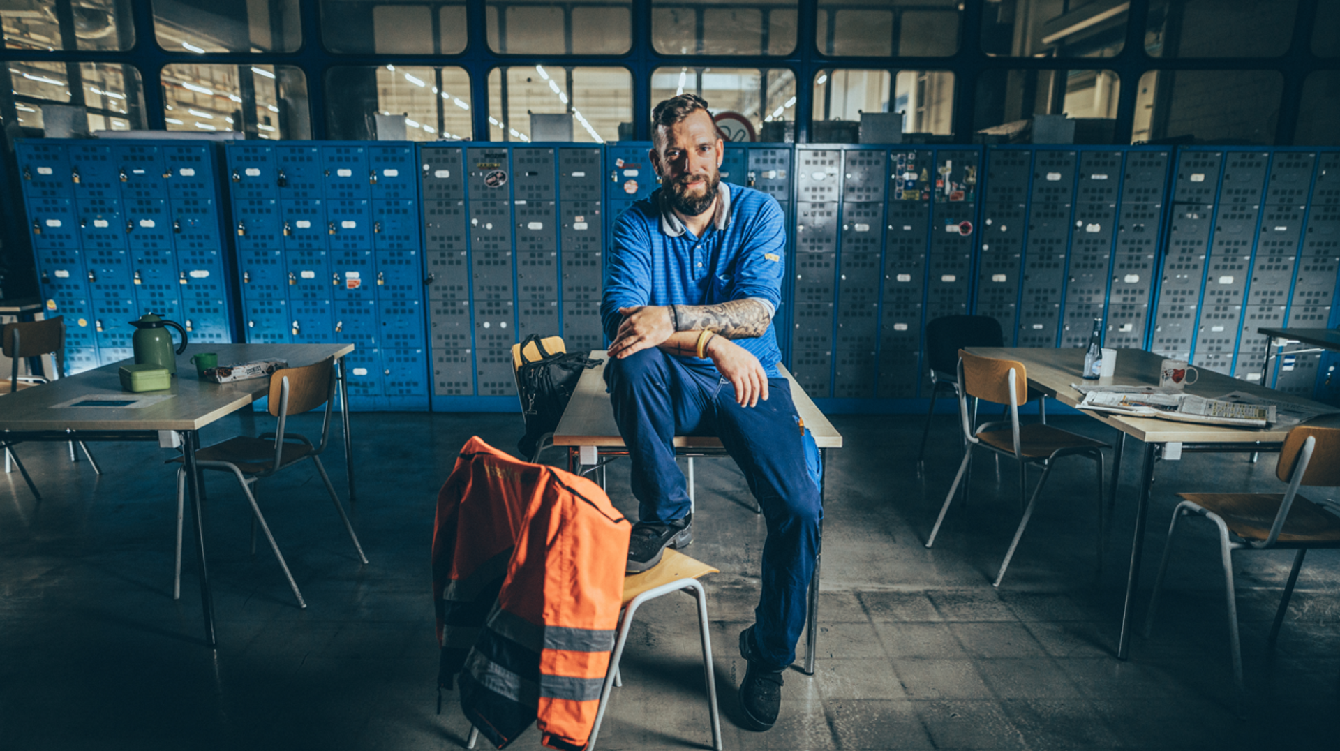 Ein Mitarbeiter in Arbeitskleidung sitzt auf einem Tisch mit einem Fuß auf dem Stuhl in einem Werkraum von thyssenkrupp. Spinde und Arbeitsplätze sind im Hintergrund zu sehen.