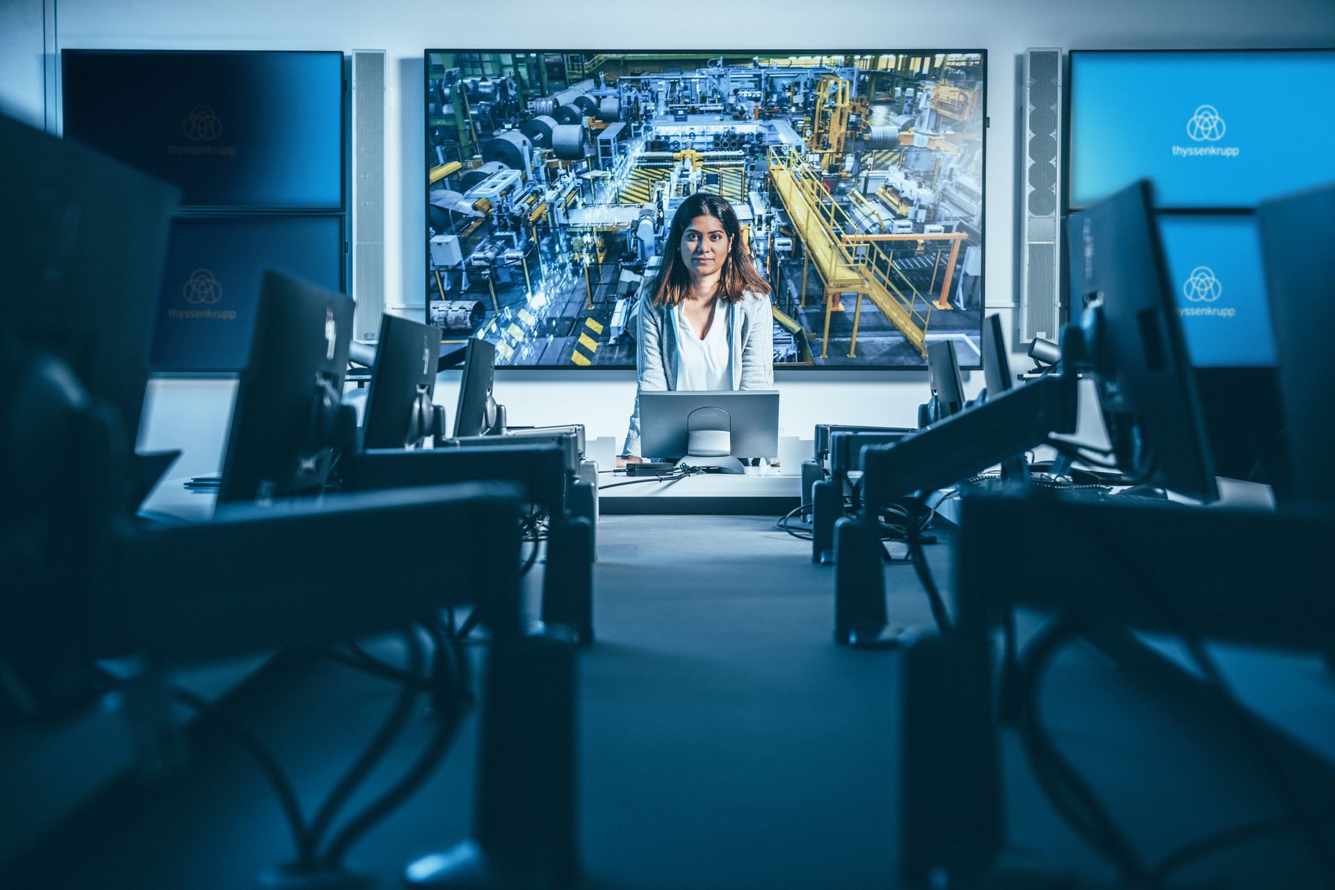 Eine Person sitzt an einem Arbeitsplatz im Kontrollraum, umgeben von mehreren Monitoren. Auf der großen Leinwand im Hintergrund ist eine industrielle Produktionsanlage zu sehen.