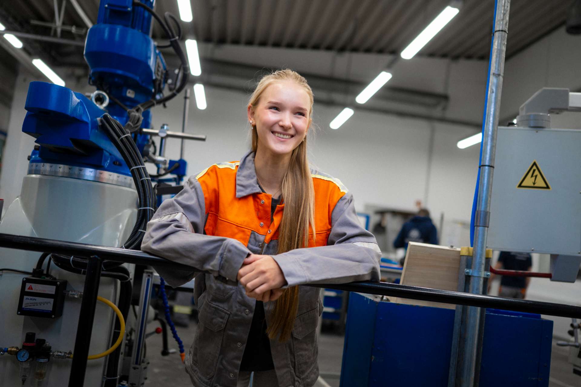 Eine Schülerin in orange-grauer Arbeitskleidung steht lächelnd in einer Werkhalle von thyssenkrupp und lehnt entspannt an einer Maschine.